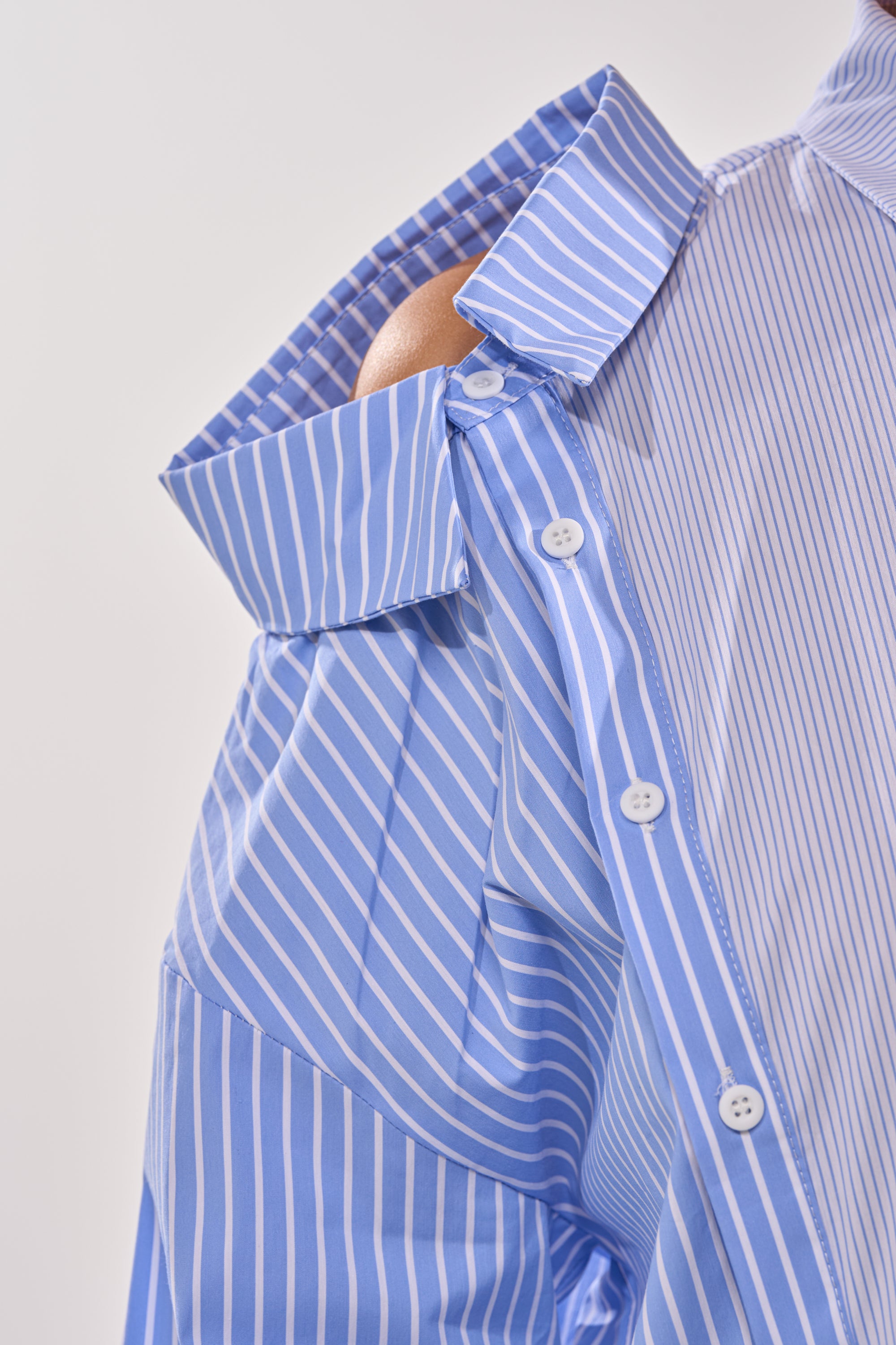 A close-up of the ALL OVER THE PLACE BUTTON DOWN, a blue and white striped blouse with white buttons. The deconstructed design exposes one shoulder, while the open collar sits against a light background. 11