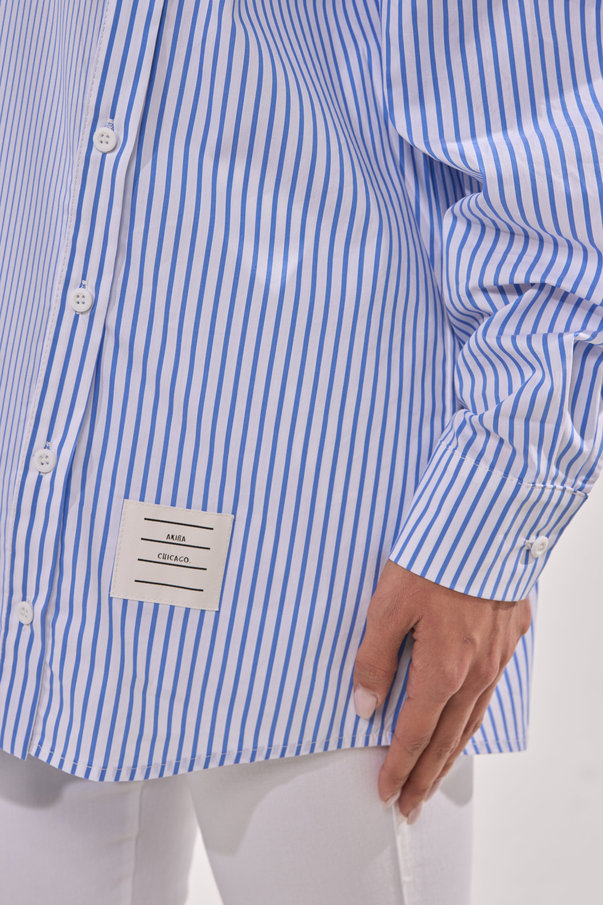 Wearing the ALL OVER THE PLACE BUTTON DOWN, a blue and white striped blouse with a hem label, paired with white pants. Their hand rests at their side. 12