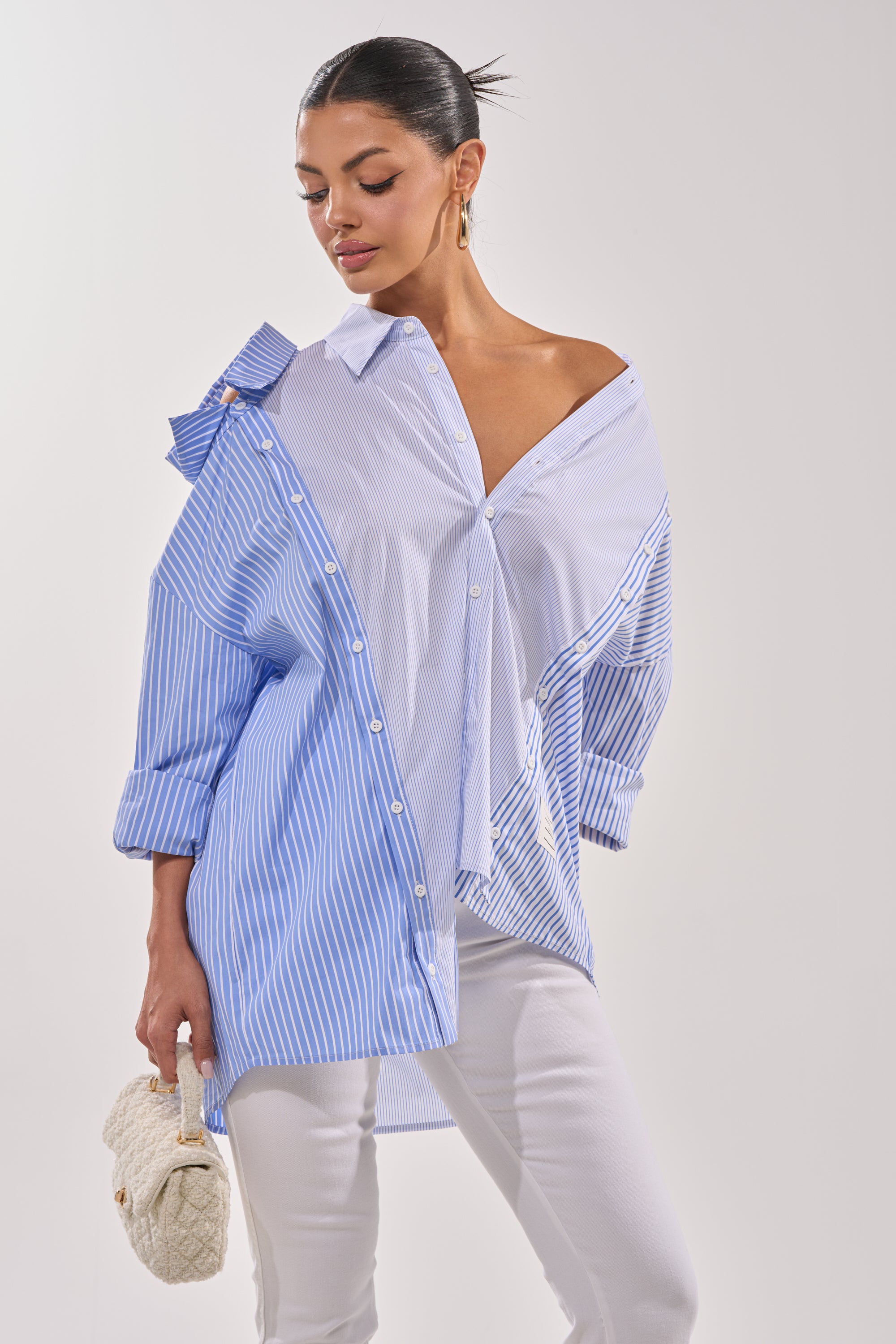 A woman wears the ALL OVER THE PLACE BUTTON DOWN, a loose, asymmetrical blue and white striped shirt, paired with white pants. She holds a white woven clutch and poses against a plain light background with her hair in a sleek bun. 2