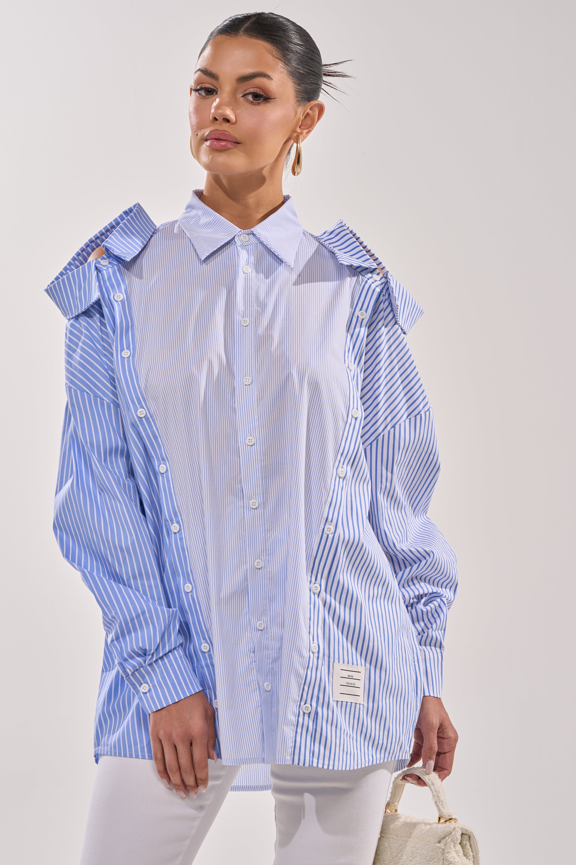 A woman wears the ALL OVER THE PLACE BUTTON DOWN, a blue and white striped blouse with cut-out shoulders, paired with white pants, gold hoop earrings, and a small beige handbag against a plain light background. 3