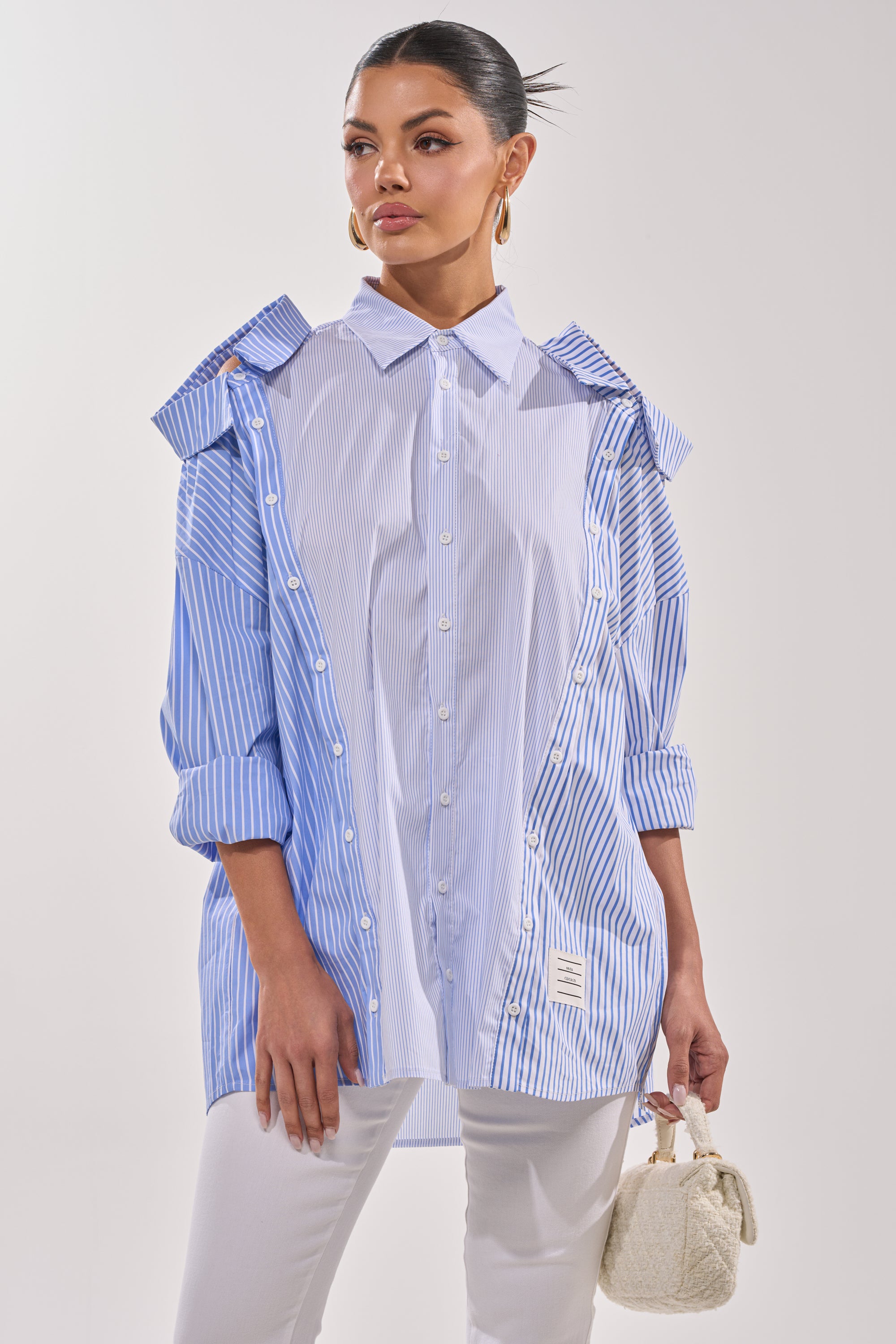 A woman wears the ALL OVER THE PLACE BUTTON DOWN—a blue and white striped blouse—over a white collared shirt, paired with white pants and gold hoop earrings. She holds a cream textured handbag against a plain background. 4