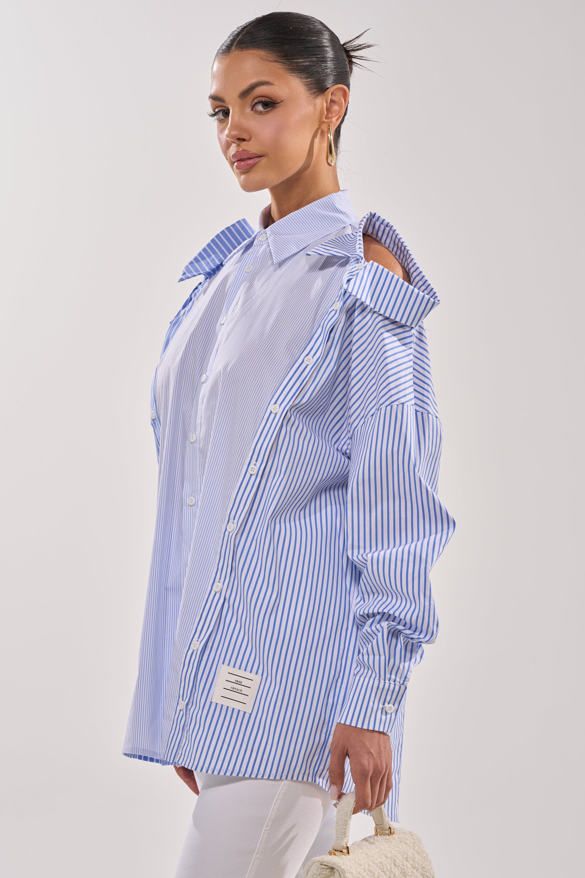 A woman wears the ALL OVER THE PLACE BUTTON DOWN, a blue and white striped blouse with cut-out shoulder details, paired with white pants, gold hoop earrings, and a woven handbag against a plain background. 5