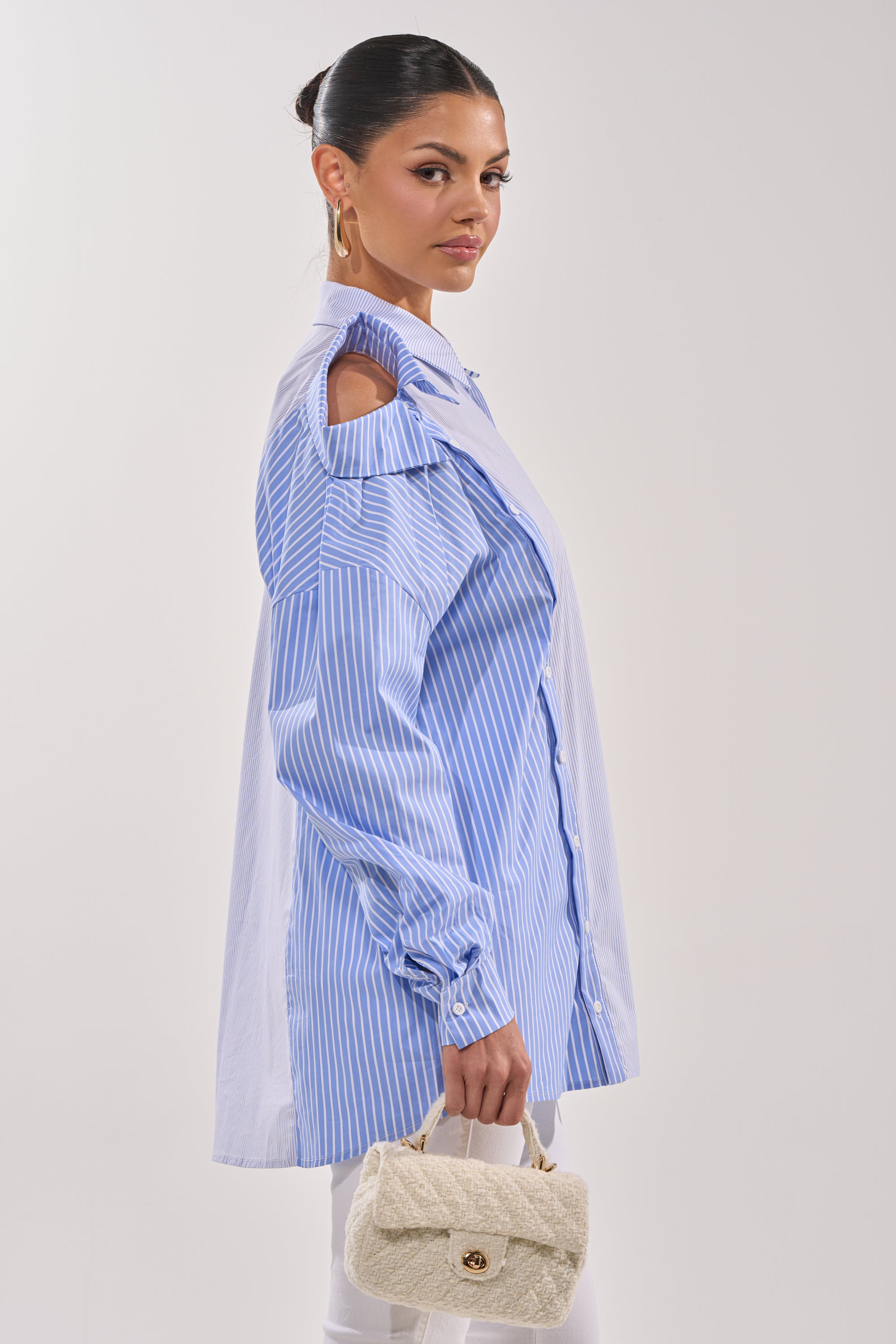 She wears the ALL OVER THE PLACE BUTTON DOWN, a blue and white striped cold-shoulder blouse, with white pants and a cream textured handbag. Her hair is tied back, she faces sideways, and finishes the look with gold hoop earrings. 6