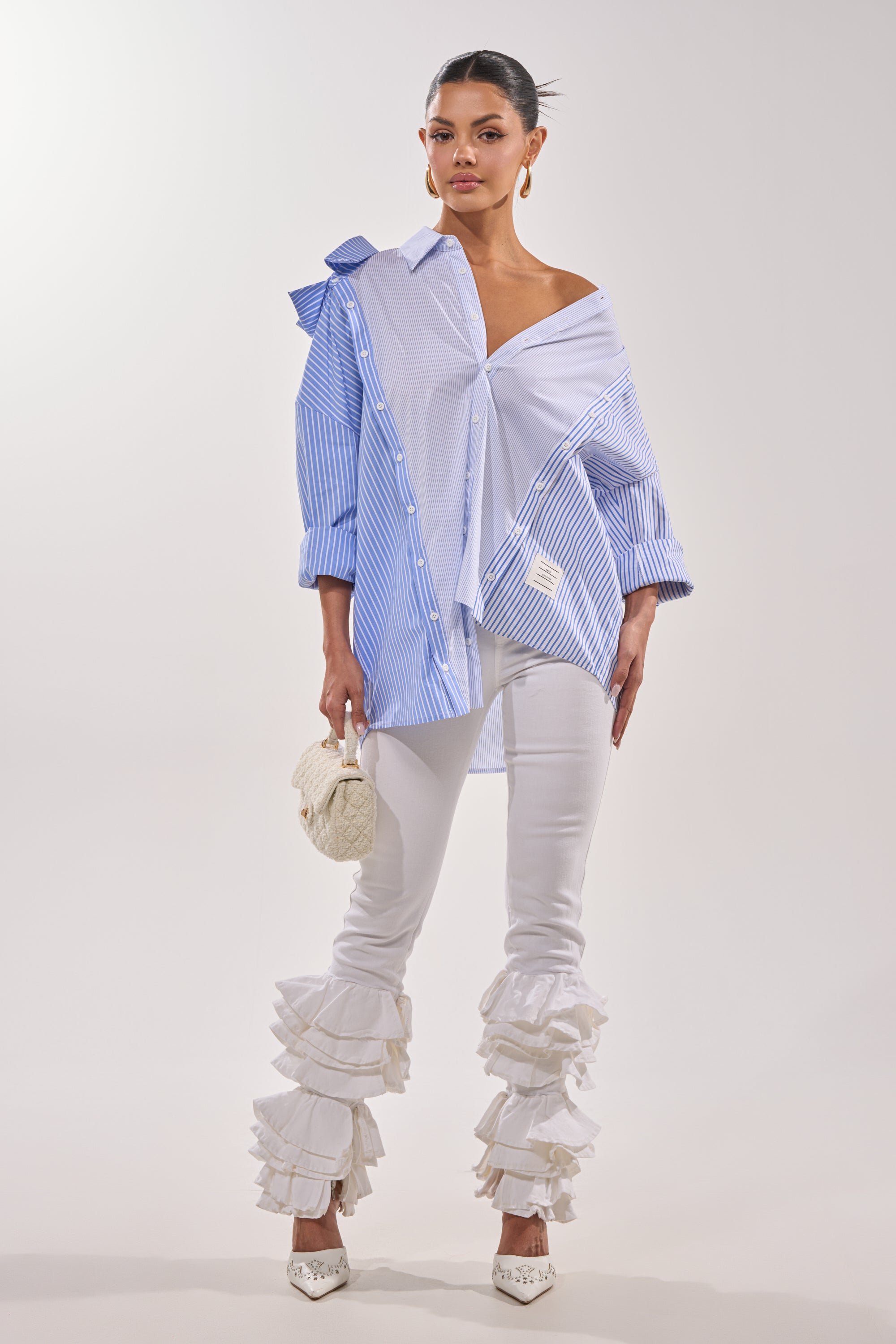 A woman poses in the ALL OVER THE PLACE BUTTON DOWN, a blue and white striped asymmetrical blouse, paired with ruffled-ankle white pants, pointed embellished heels, a textured clutch, slicked-back hair, and hoop earrings. 8