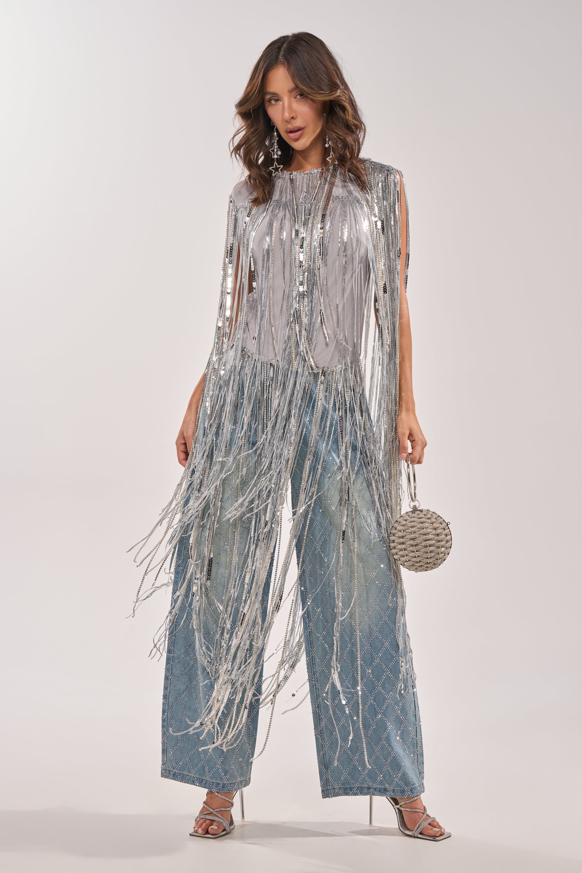 A woman poses in the DRIP CITY FRINGE TOP, a silver sequin statement piece with long metallic tassels, styled over wide-leg blue jeans. She completes her party look with a round woven handbag and silver high-heeled sandals. 0