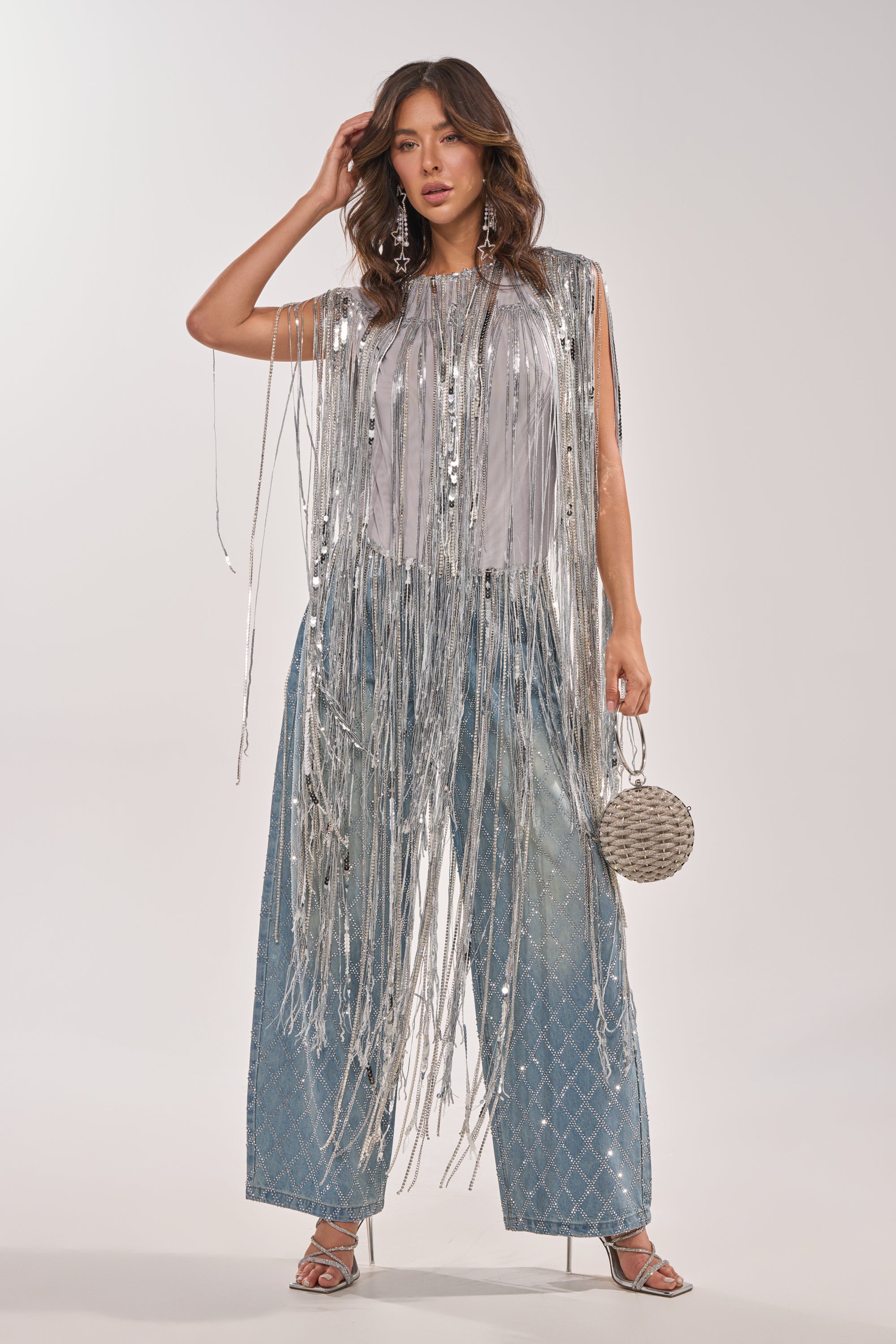A woman models the DRIP CITY FRINGE TOP layered over a white shirt, styled with wide-leg diamond-pattern blue jeans, silver heels, and a round silver handbag—her wavy hair and confidence complete the ultimate statement clubwear look. 1