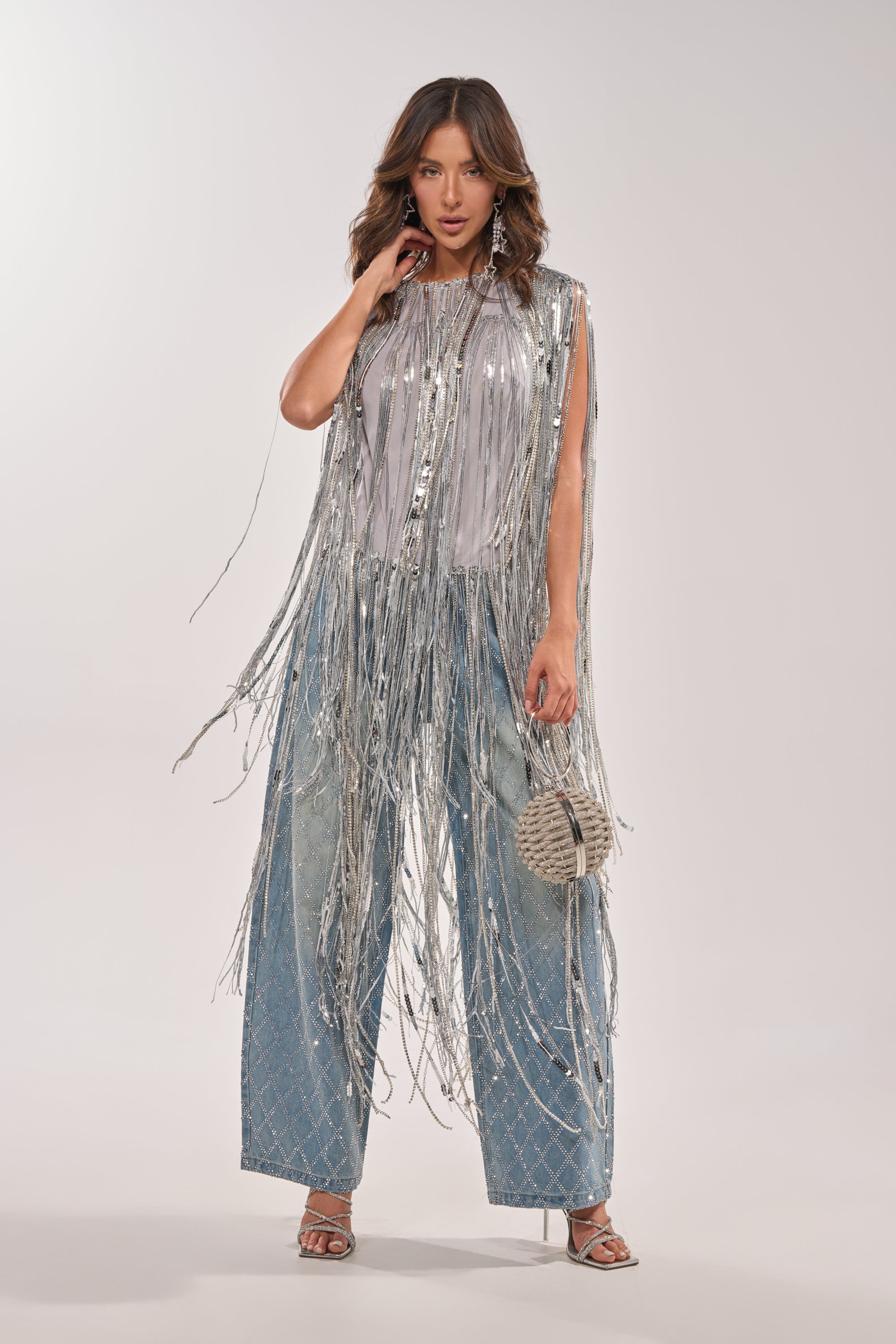 A woman poses in the DRIP CITY FRINGE TOP over a white shirt, paired with wide-leg blue crisscross jeans, silver heels, and a round silver textured handbag—a bold clubwear party look against a plain background. 2