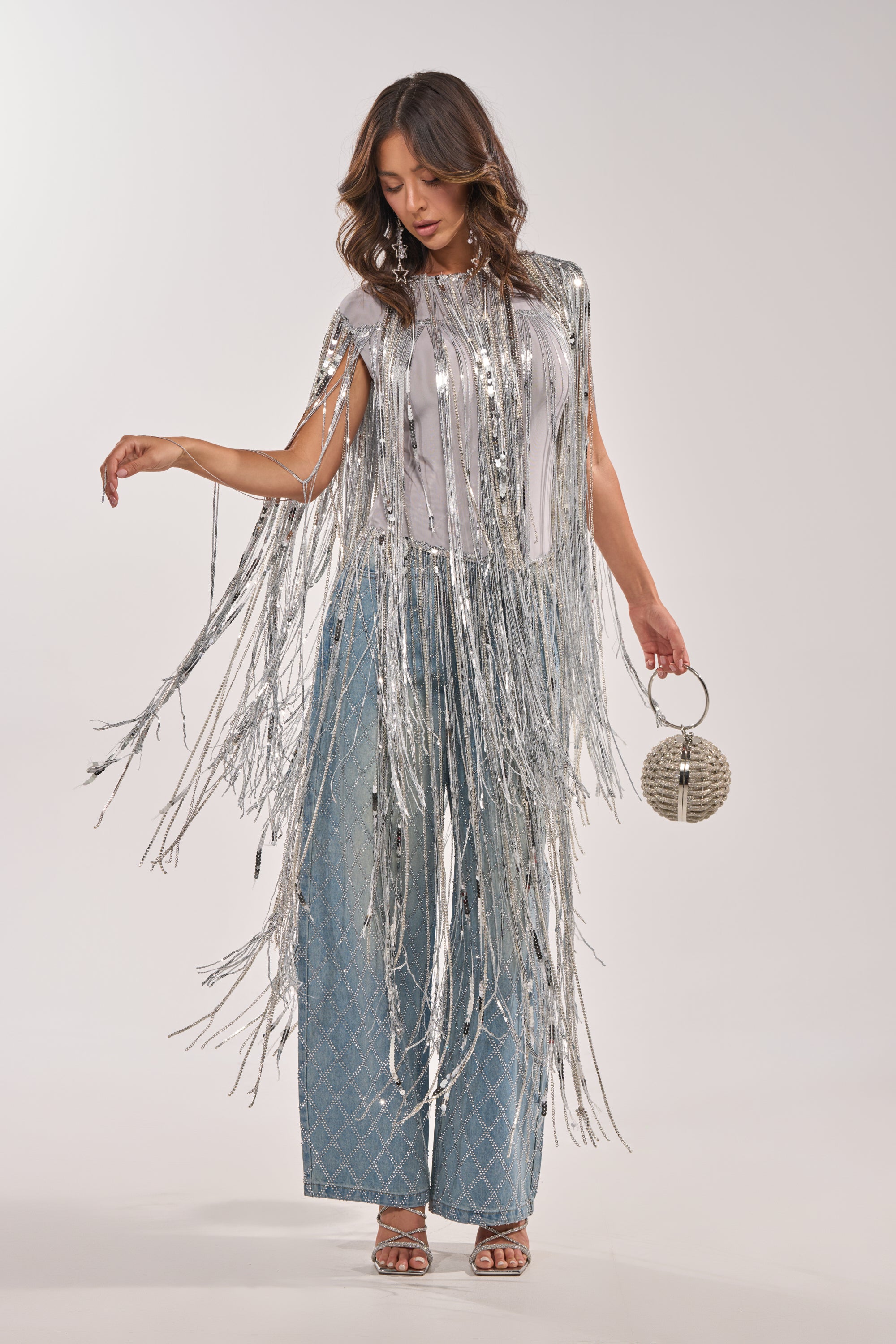 A woman models the DRIP CITY FRINGE TOP with light blue patterned wide-leg pants and silver heels, posing with a round silver handbag. Her loose, wavy hair adds flair to this chic party look against a plain, light background. 3