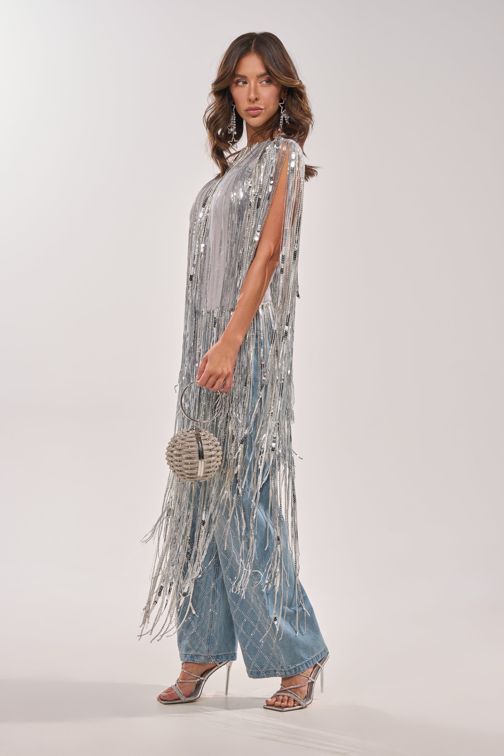 A woman poses against a plain light background in the DRIP CITY FRINGE TOP—a silver sequin fringe piece—paired with wide-leg blue jeans, strappy high heels, and a round silver bag. 4