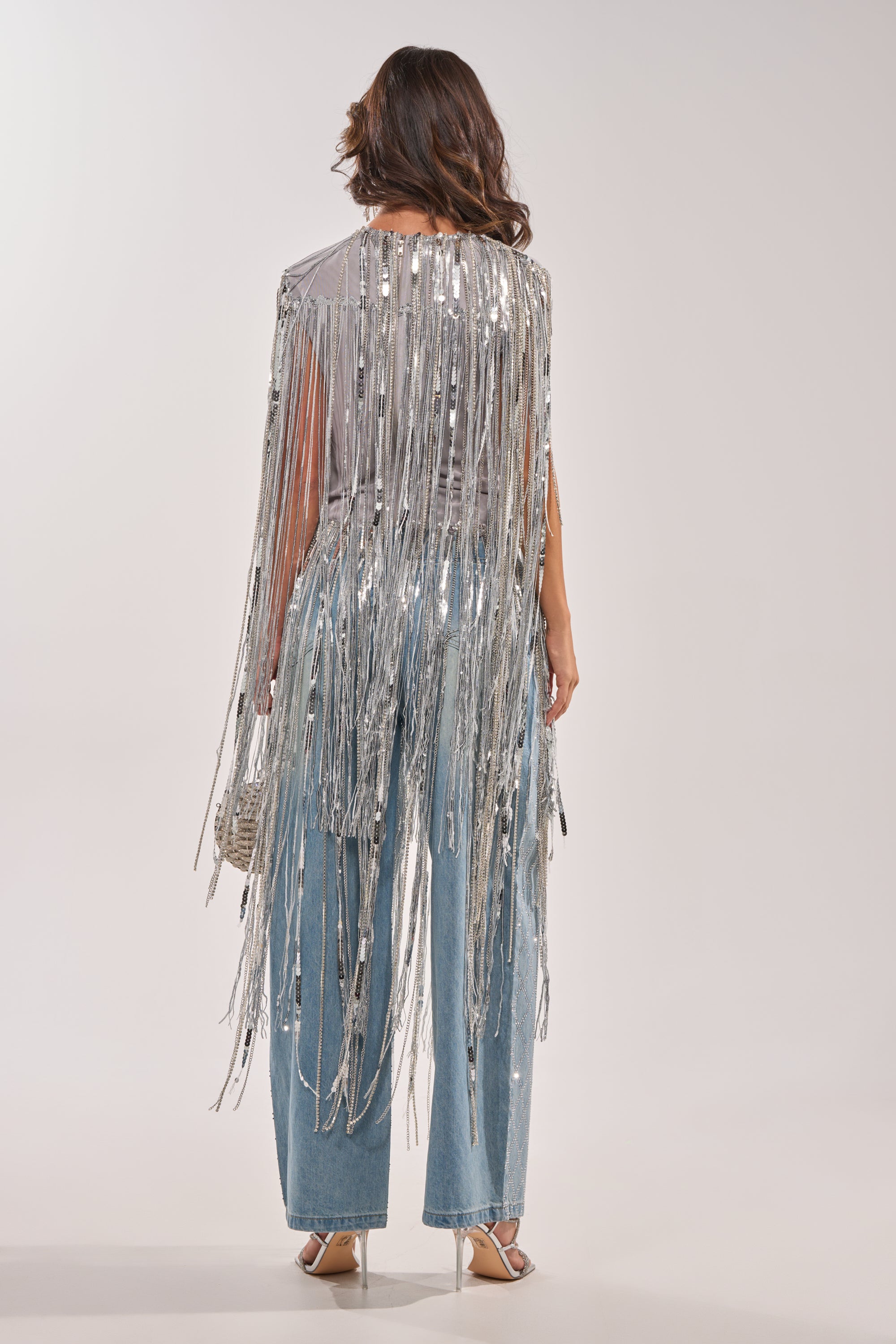 A woman faces away, wearing the DRIP CITY FRINGE TOP—a silver sequin fringe style—over light blue jeans and silver heels. She carries a small silver handbag, her wavy hair loose, styled effortlessly against a plain white background. 5