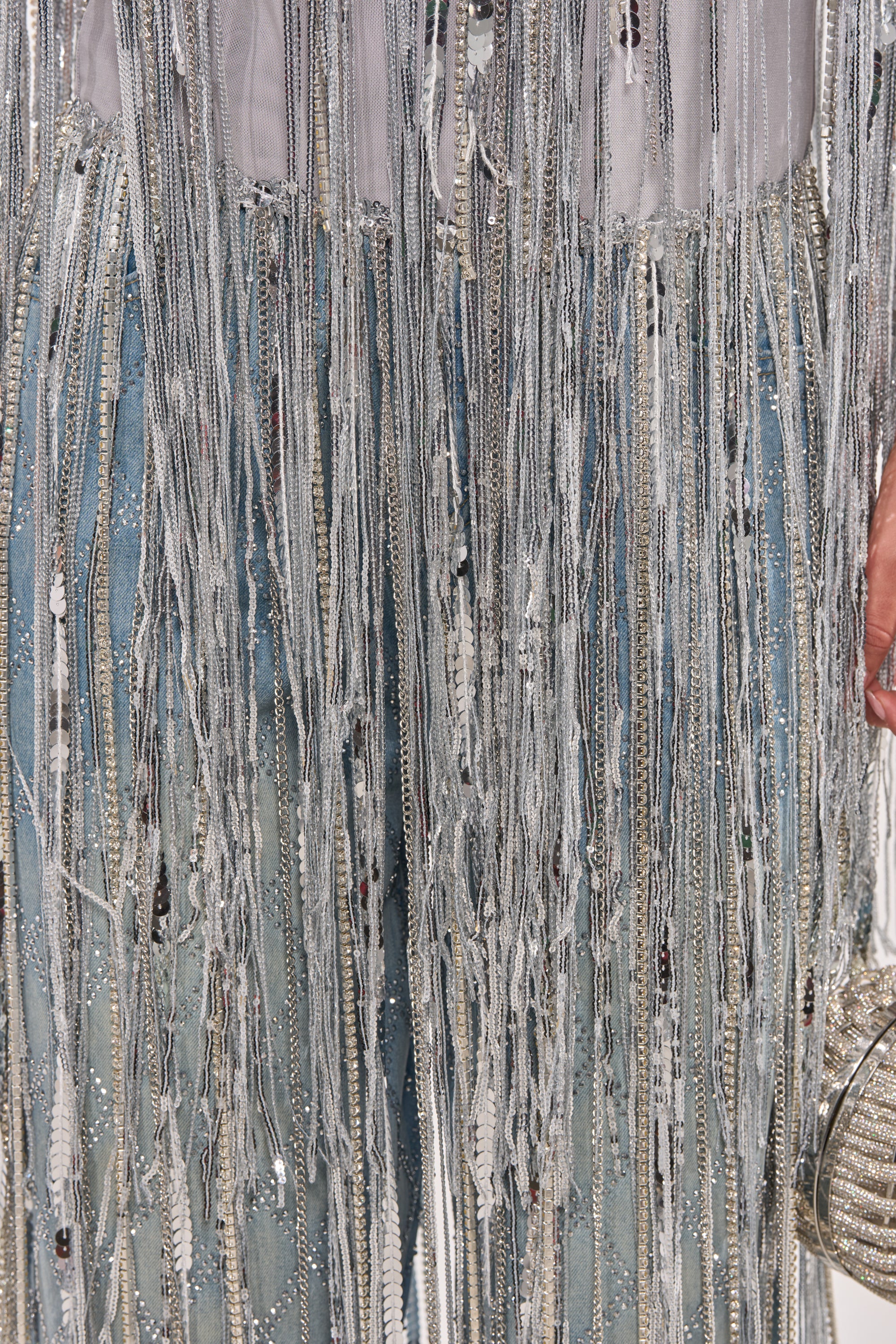 Close-up of a person wearing the DRIP CITY FRINGE TOP styled with blue jeans featuring long, silver-beaded fringe and white fabric strips. They accessorize with a small round metallic handbag for a chic party look. 7