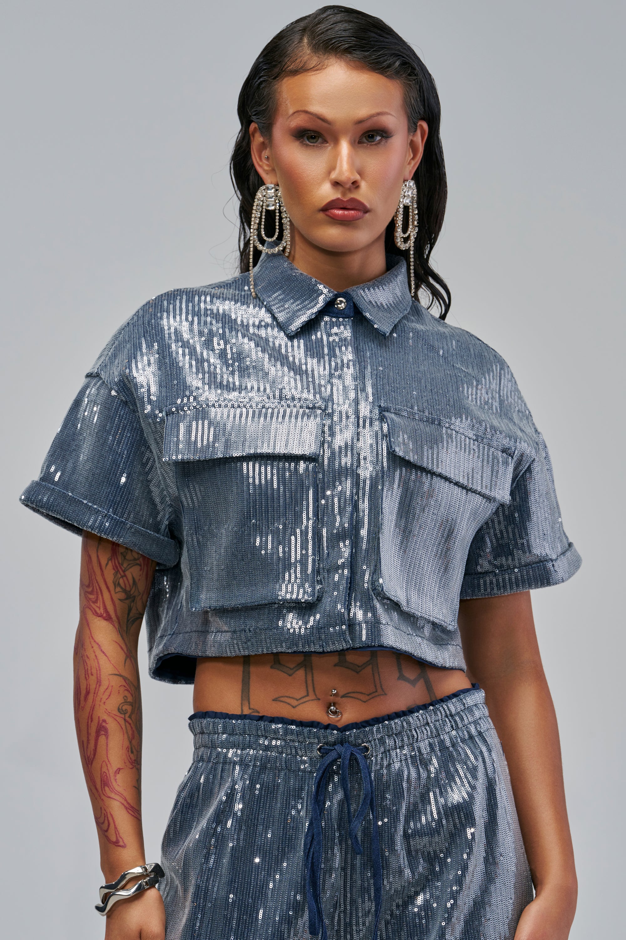 A model wearing a SHAKE IT GIRL SEQUIN BUTTON DOWN: a short-sleeved, cropped, navy sequin top designed with a button-down front and two chest pockets.