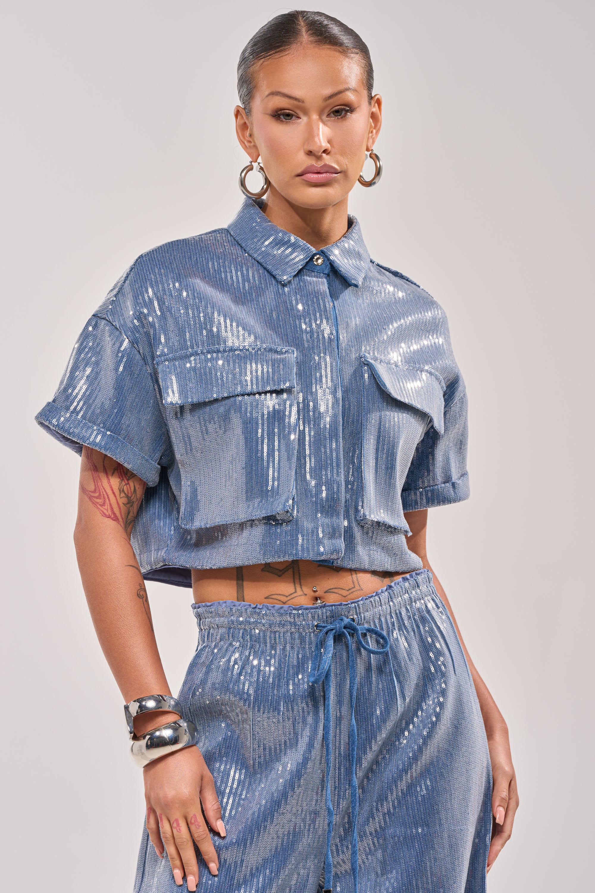 A woman exudes confidence in the SHAKE IT GIRL SEQUIN BUTTON DOWN—a shiny blue shirt with chest pockets—paired with matching drawstring pants, hoop earrings, and bracelets. Her bold look stands out against a plain background. 1