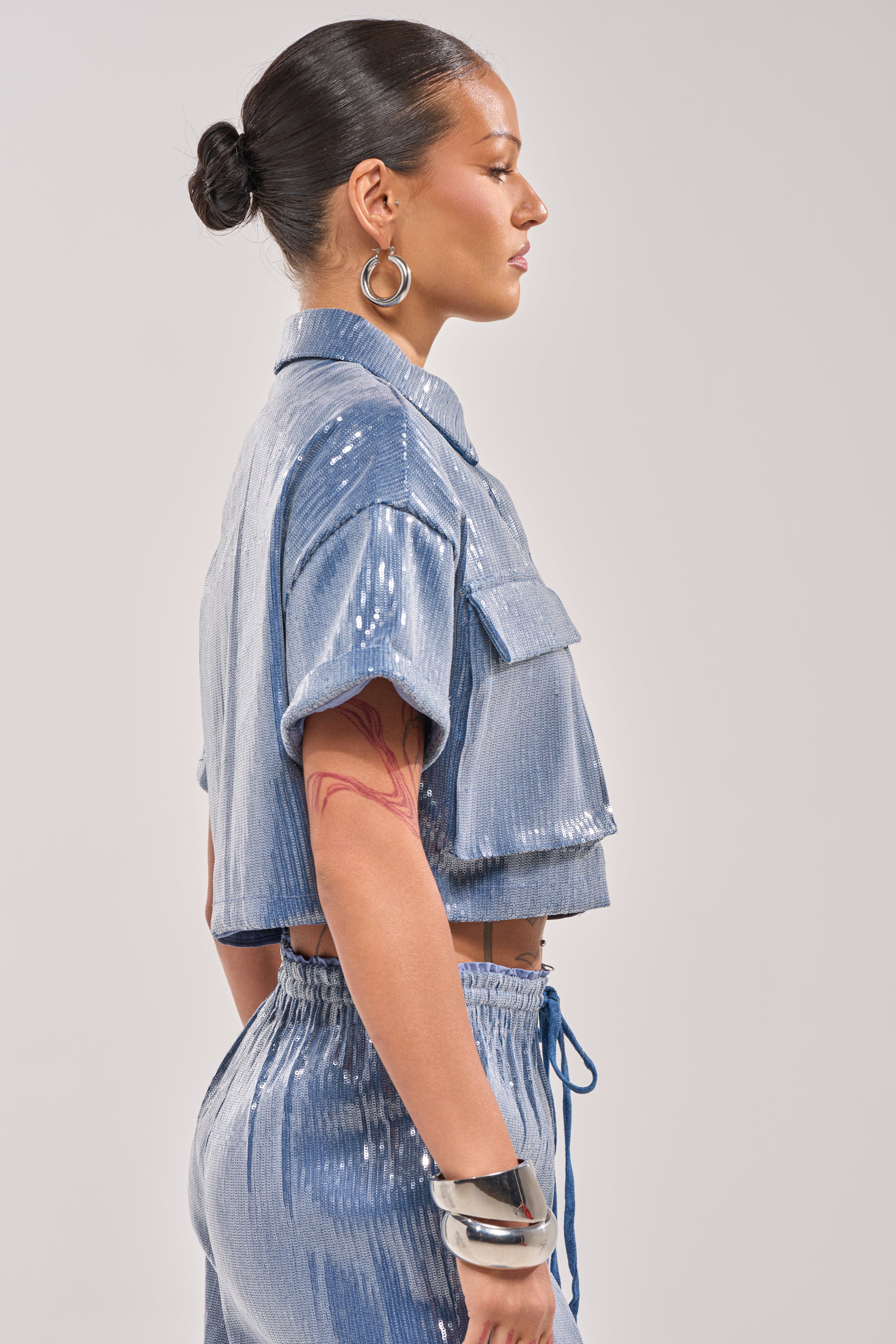 A woman with dark hair in a bun poses in profile against a plain background, wearing elevated casual style: the shiny blue SHAKE IT GIRL SEQUIN BUTTON DOWN and matching pants. She accessorizes with hoop earrings, a silver cuff bracelet, and shows off an arm tattoo. 3