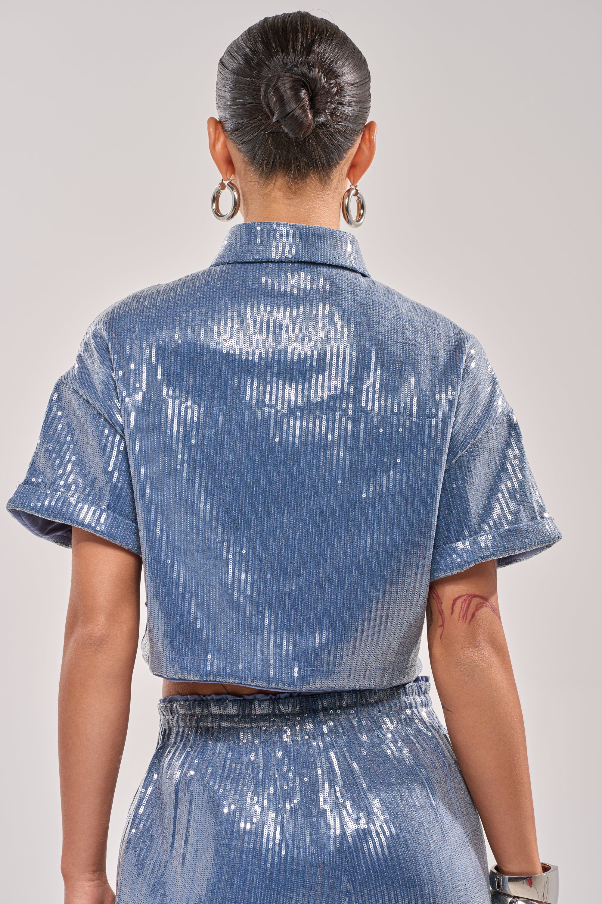 A person with dark hair in a sleek bun is seen from the back, styled casually yet chic in the SHAKE IT GIRL SEQUIN BUTTON DOWN and matching pants. Hoop earrings, a bracelet, and an arm tattoo complete this standout look. 4