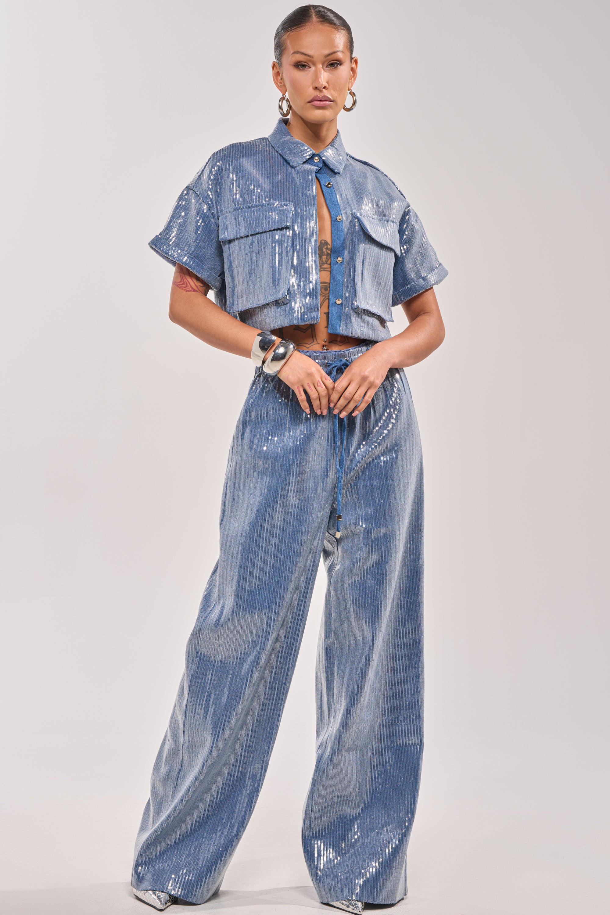 A woman stands confidently in the SHAKE IT GIRL SEQUIN BUTTON DOWN, a shiny light blue cropped shirt with large front pockets and matching wide-leg pants. Hoop earrings, bracelets, and slicked-back hair complete her chic look. 5