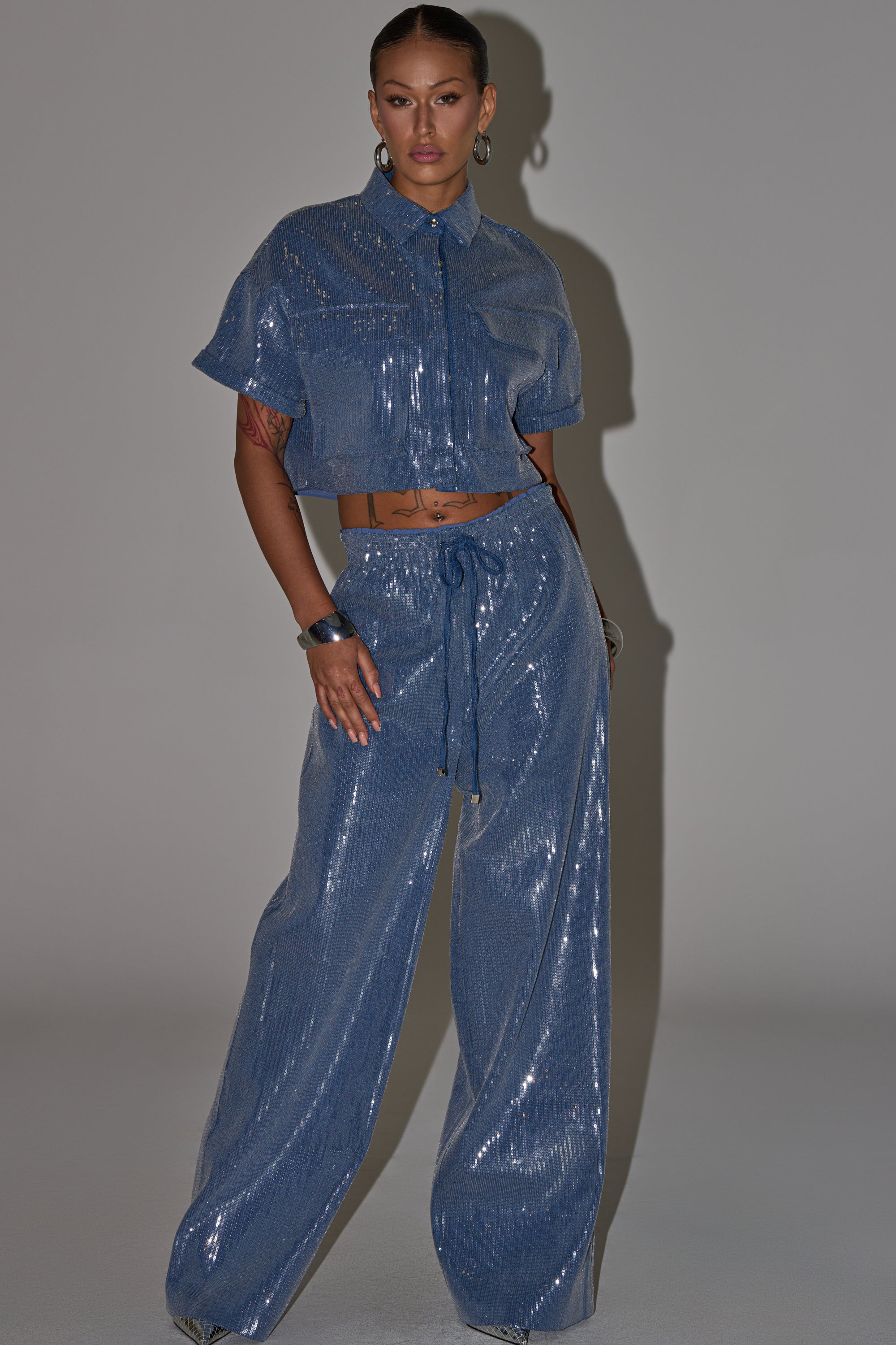 A person stands confidently in the SHAKE IT GIRL SEQUIN BUTTON DOWN, a shiny blue cropped shirt paired with matching wide-leg pants. This sparkly, light-reflective outfit is perfect for elevated casual styling with hoop earrings and slicked-back hair. 6