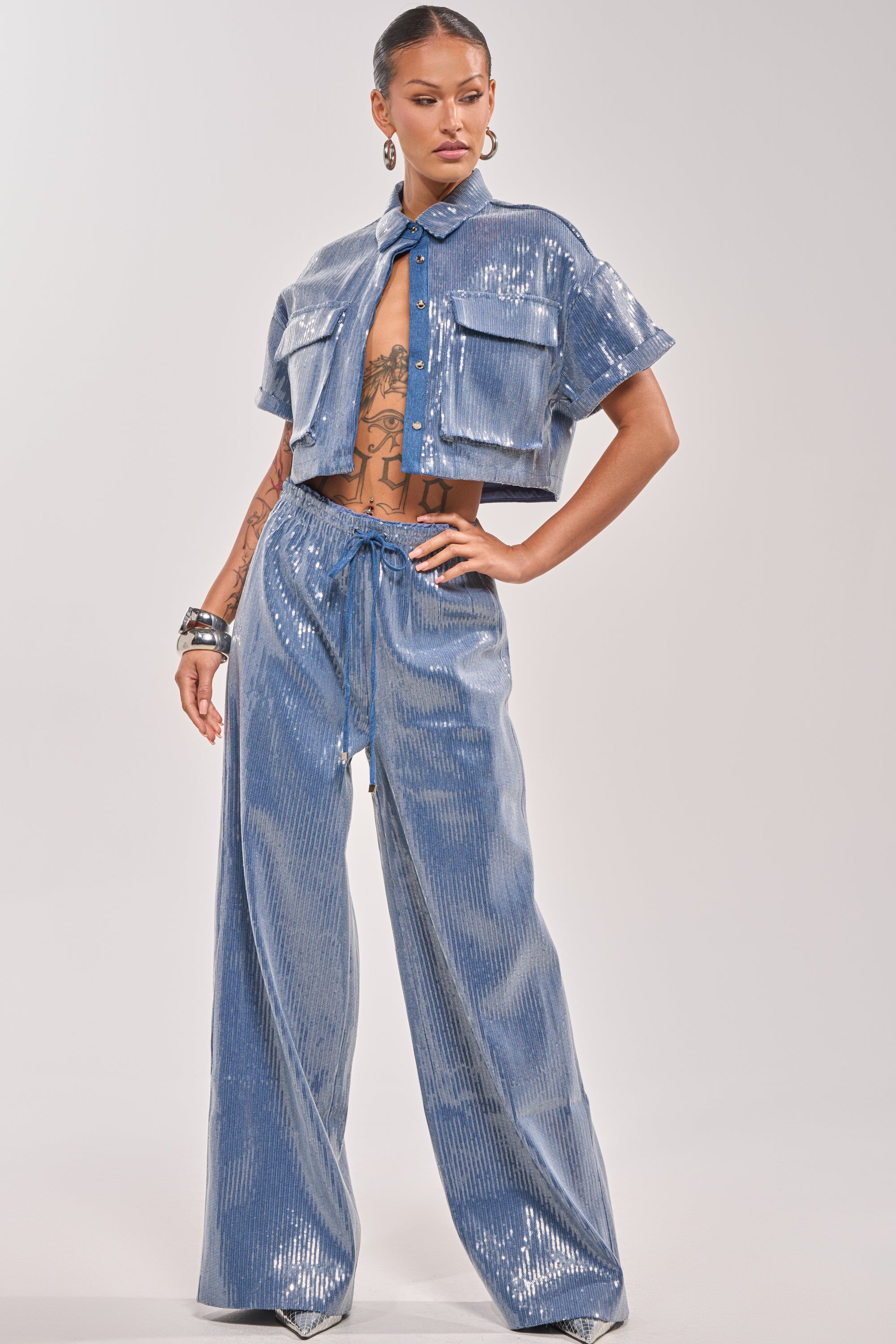 A woman poses confidently in the SHAKE IT GIRL SEQUIN BUTTON DOWN, a shiny light blue cropped sequin shirt, paired with wide-leg high-waisted pants, accessorized with hoop earrings and bracelets, exuding elevated casual style. 7