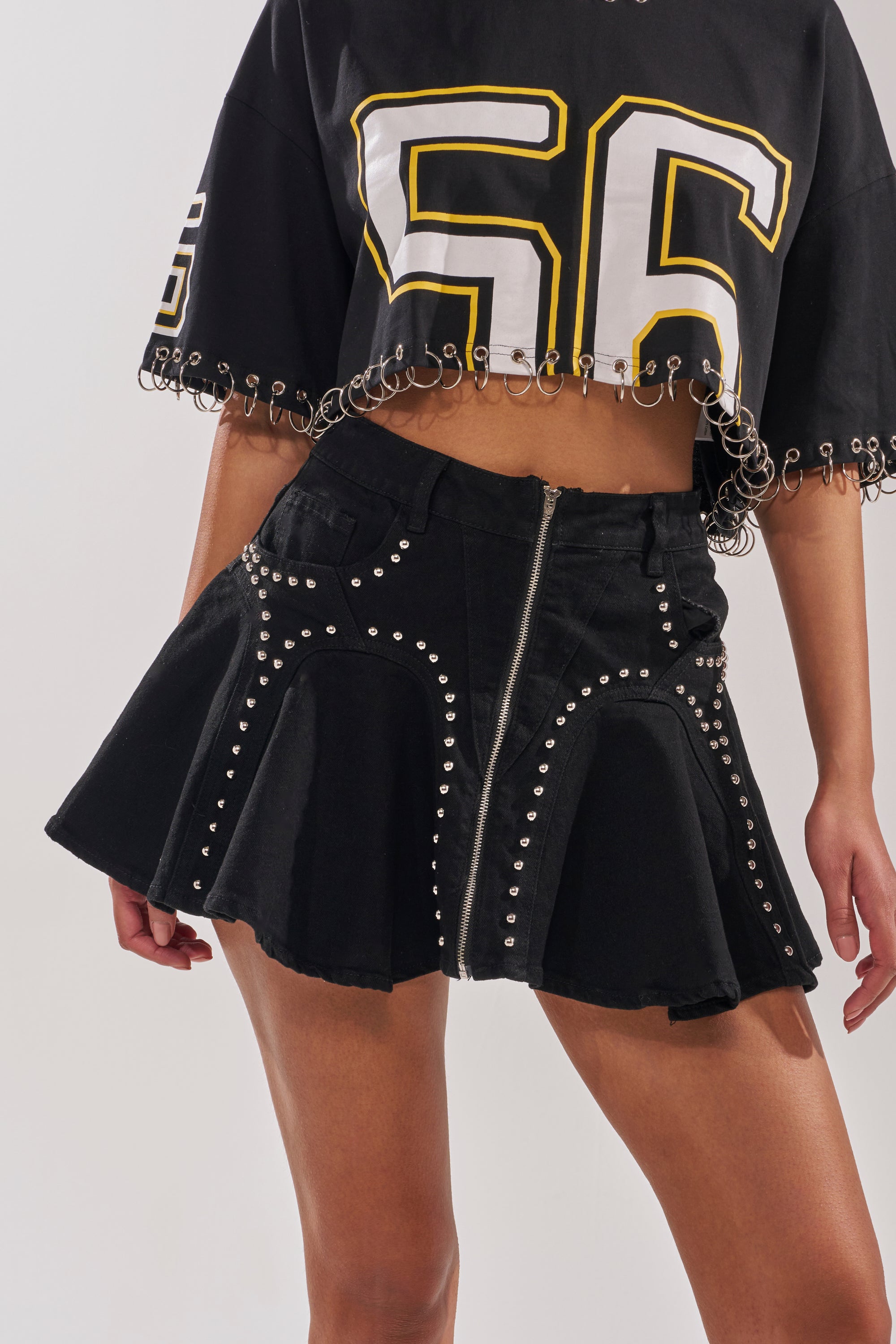 A person wearing a cropped black sports jersey with the number 56 and the LUCKY ONE DENIM MINI SKIRT, featuring studs, a flared high waist, and front zipper, stands against a plain background. 1