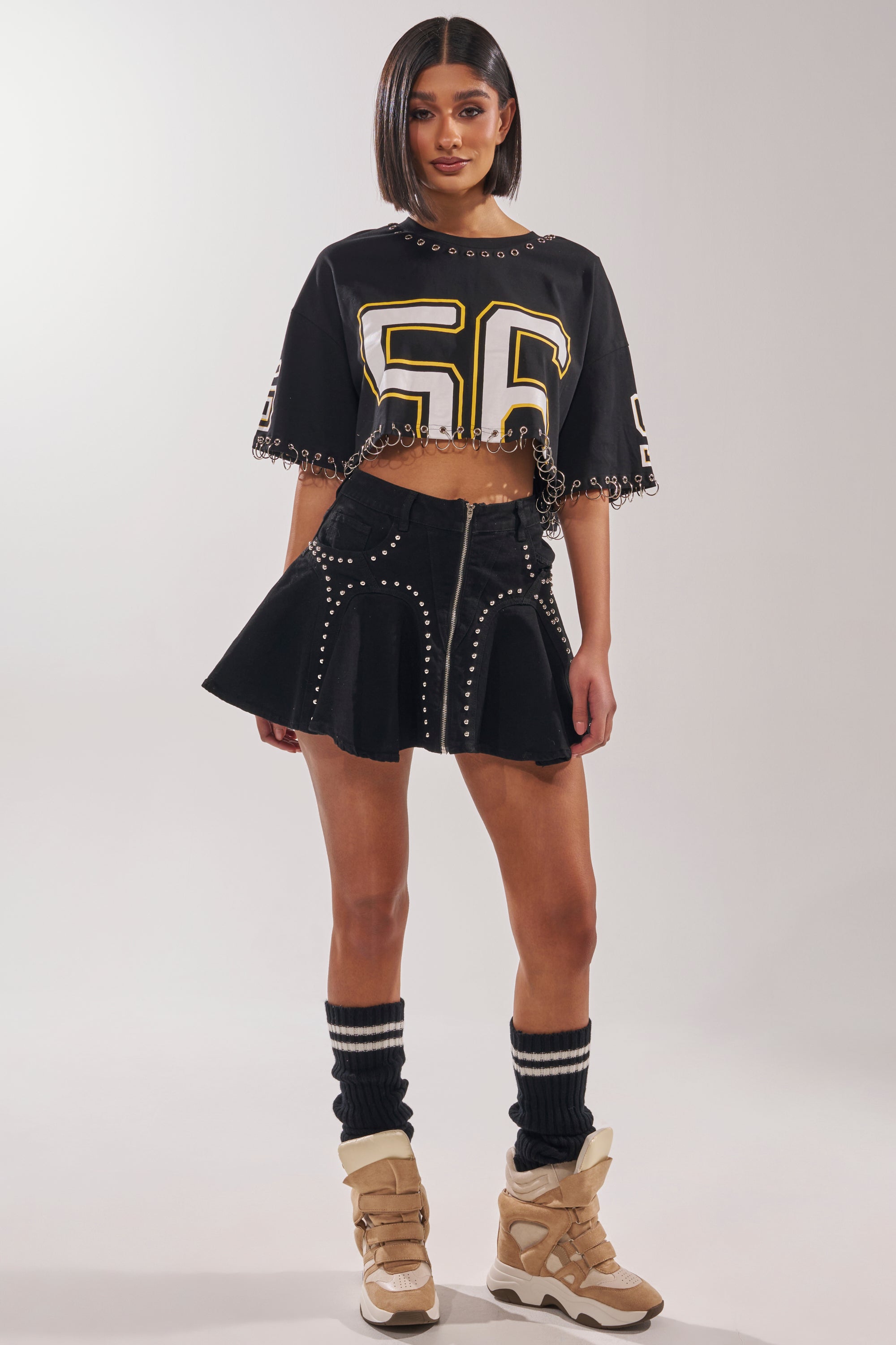 A woman stands against a plain light background wearing a black crop top with the number 56, black leg warmers, beige sneakers, and the LUCKY ONE DENIM MINI SKIRT featuring silver studs and a high-rise waist. 5