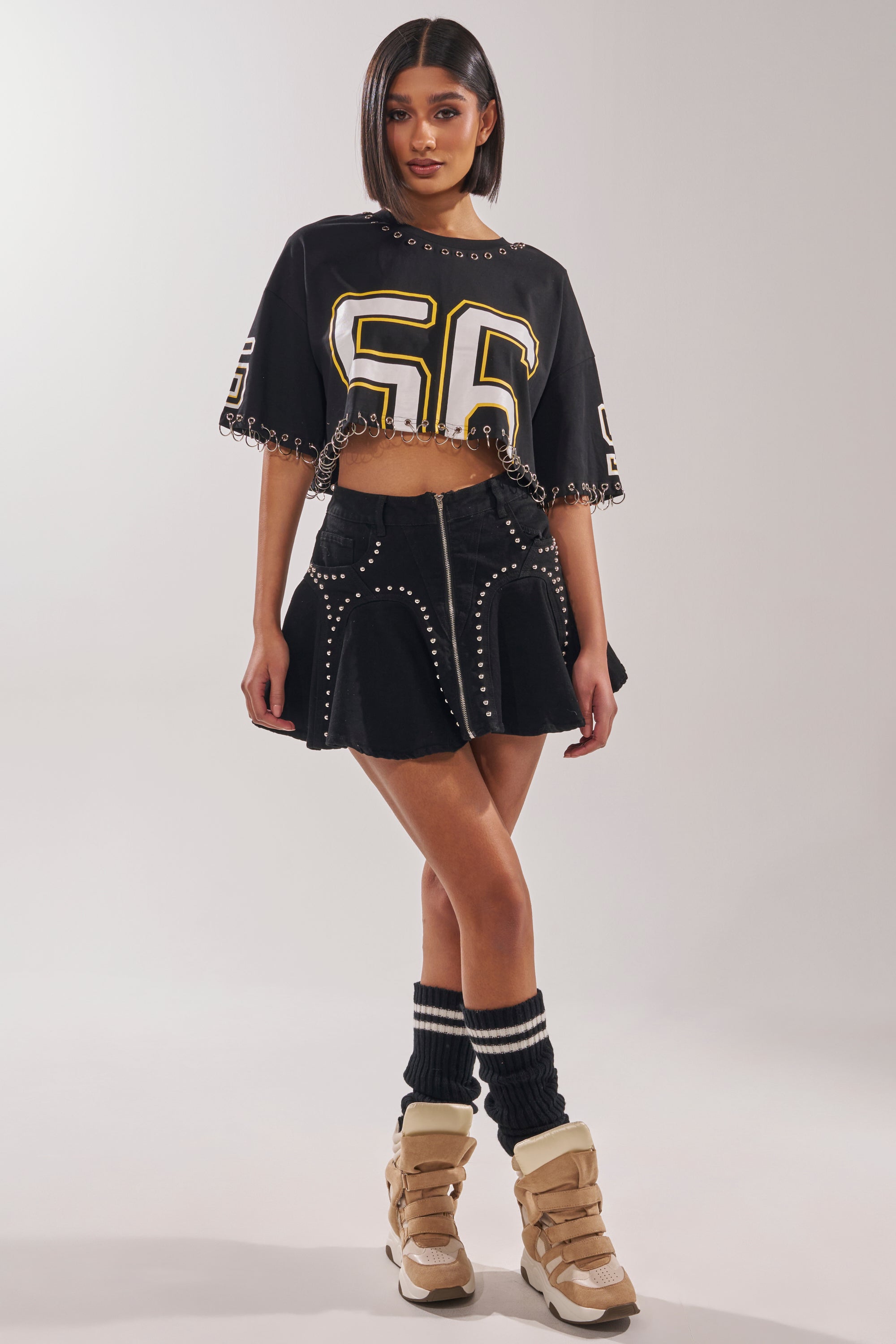 A woman wears a black crop top with "56", the LUCKY ONE DENIM MINI SKIRT featuring silver studs and a high-rise waist, black leg warmers, and beige high-top sneakers. Her hair is straight and parted in the middle. 6