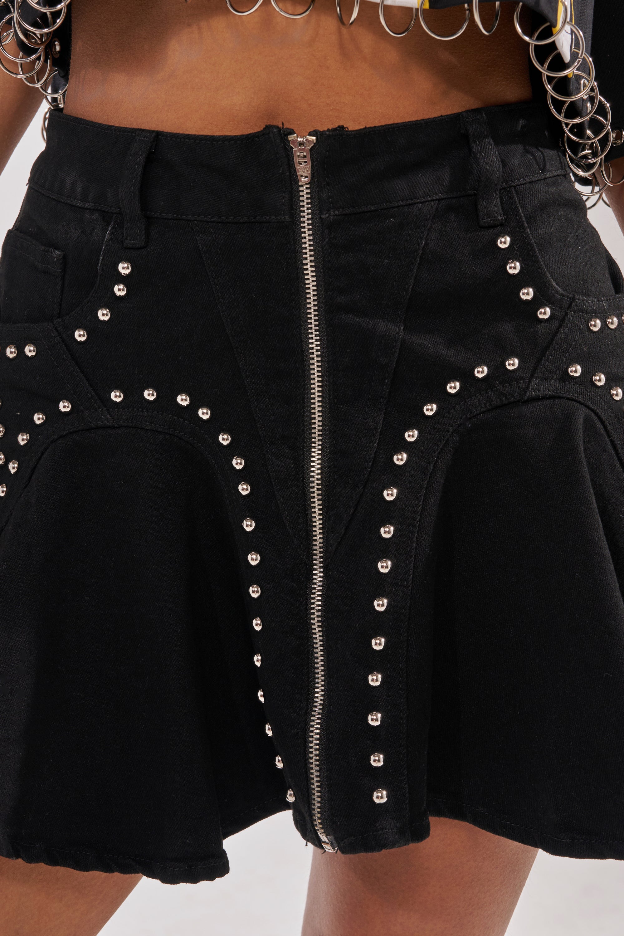 Close-up of the LUCKY ONE DENIM MINI SKIRT in black, featuring a high-rise waist, front zipper, silver stud curved embellishments, belt loops, and paired with a chain-accented top. 7