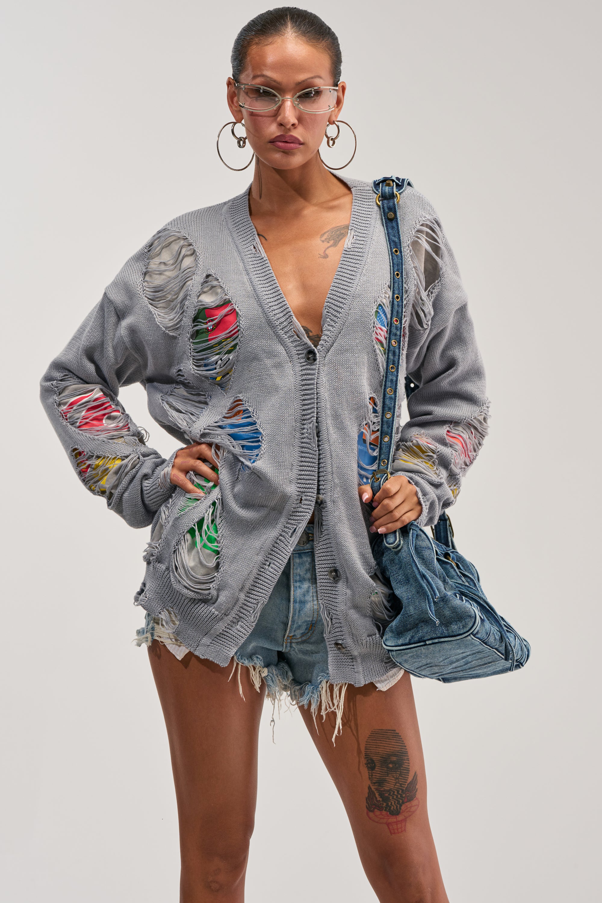 A woman styles the LYDIA DISTRESSED GRAPHIC SWEATER in grey over shorts and carries a blue denim bag. She accessorizes with large hoop earrings, clear glasses, and showcases a prominent thigh tattoo against a plain, light-colored background.