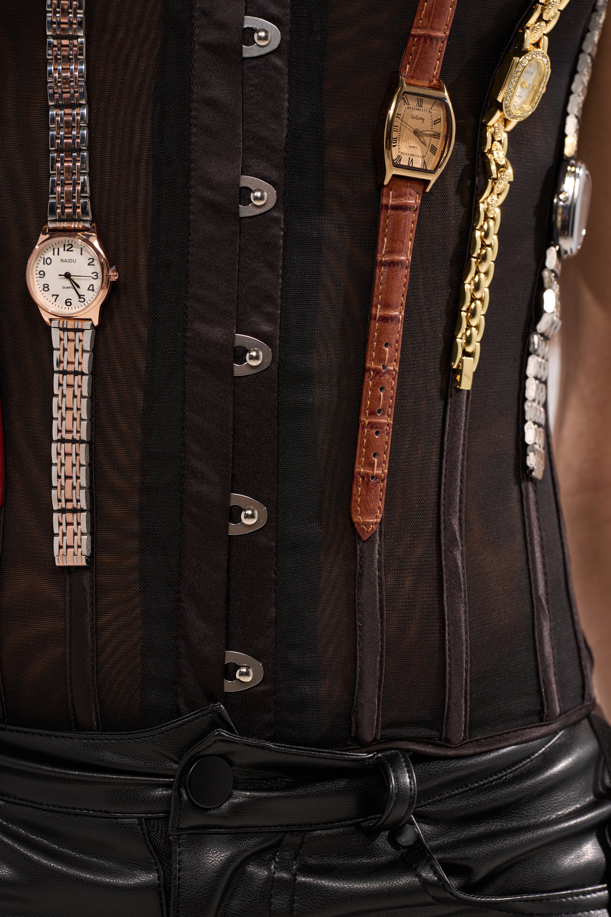 A person wears the WHAT TIME IS IT CORSET—upcycled fashion featuring multiple wristwatches and watch bands draped across the chest—styled with black leather pants and a sheer black top. 10