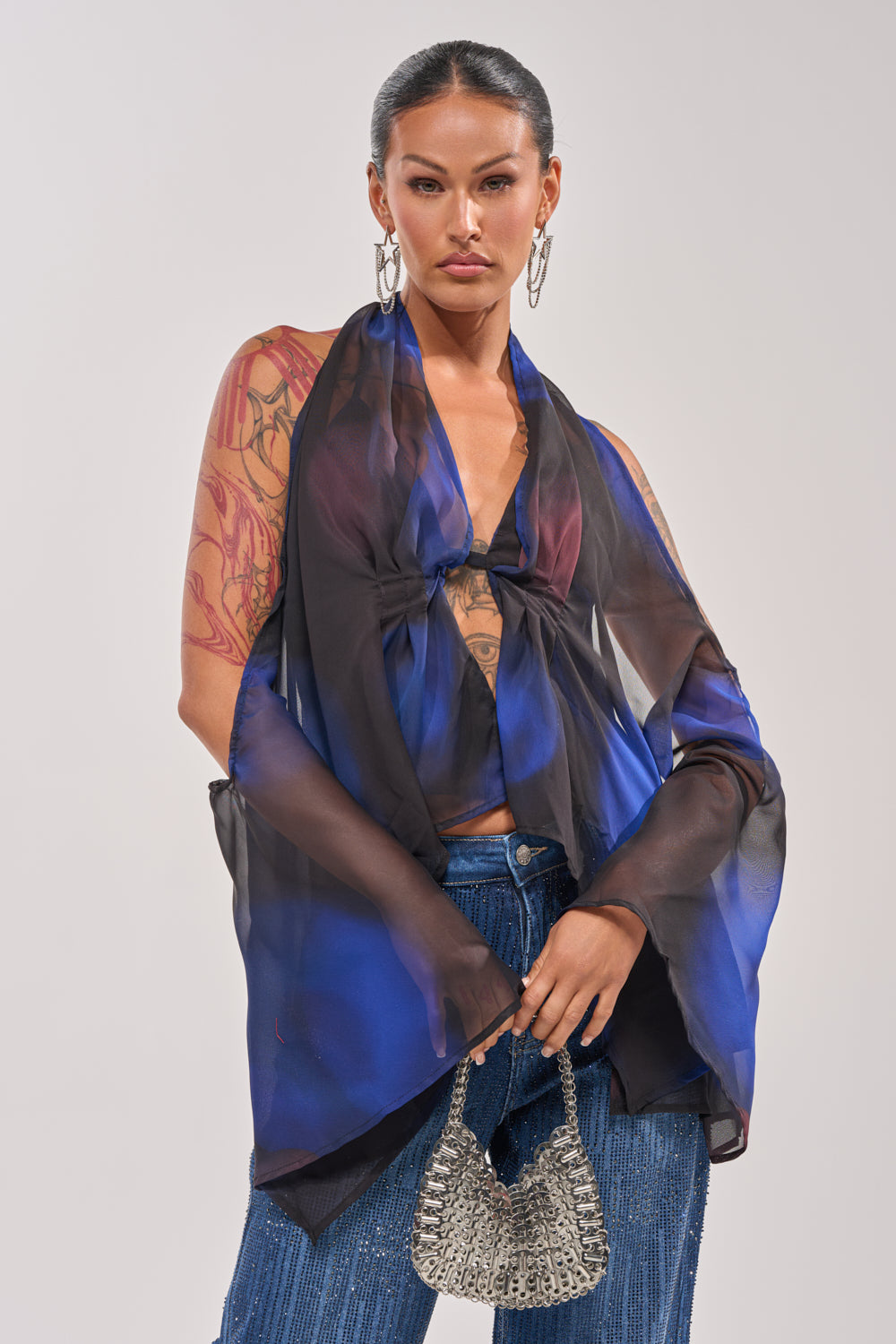 A confident woman with tattoos poses against a plain background in the FLY AWAY TIE FRONT BLOUSE, blue pants, silver hoop earrings, and a small silver handbag, perfectly showcasing vacation fashion. 0