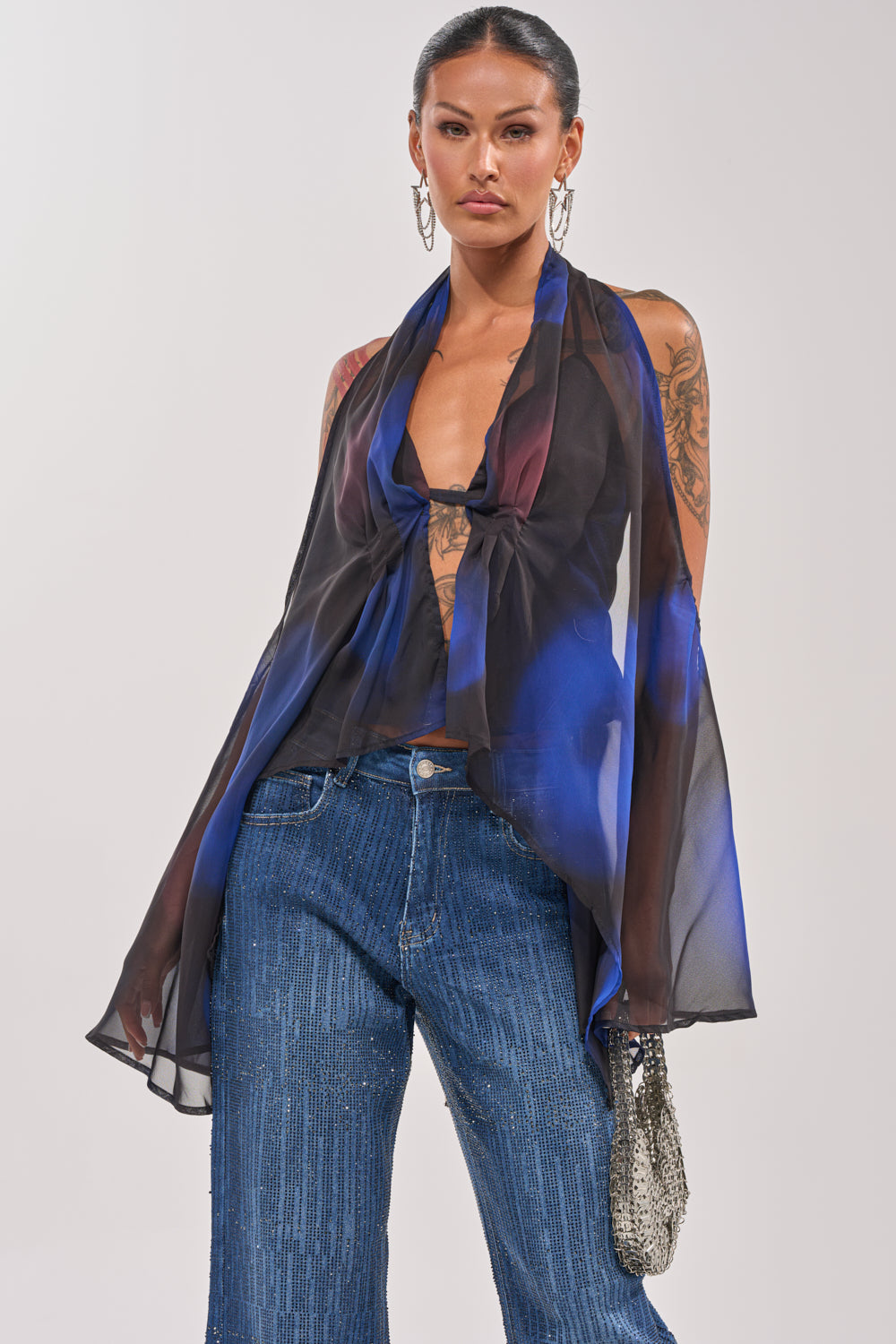 A woman wears the FLY AWAY TIE FRONT BLOUSE—a sheer, flowing blue marbled chiffon top—over blue jeans, styled with large hoop earrings, a small metallic bag, slicked-back hair, and arm tattoos. 1