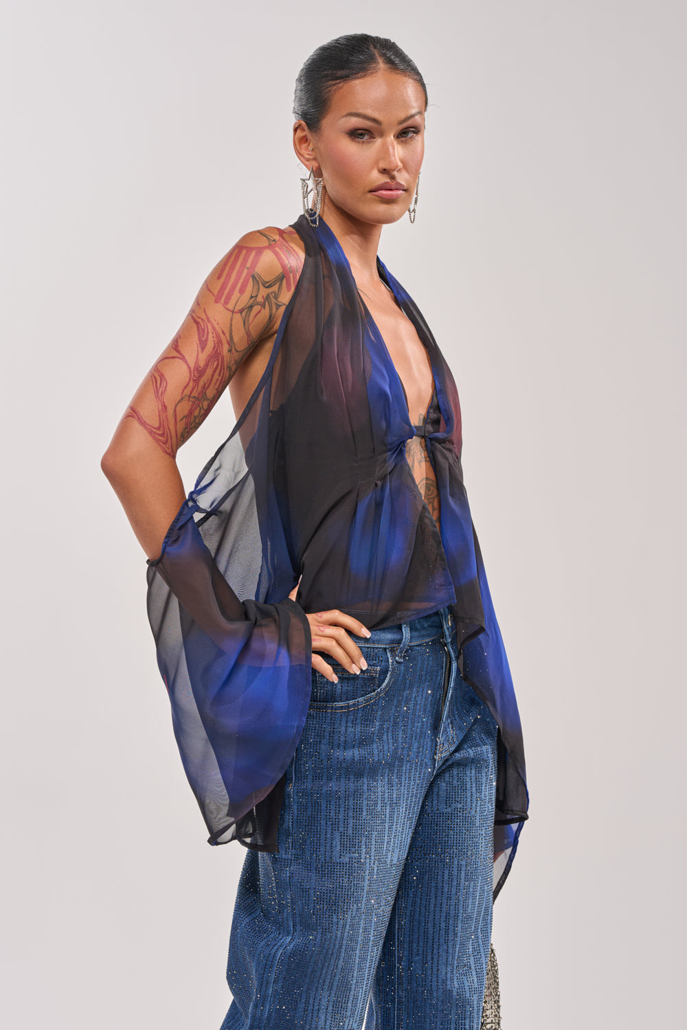 A woman with a tattooed arm wears the FLY AWAY TIE FRONT BLOUSE—a sheer, blue marbled chiffon top styled over blue jeans. With hoop earrings and her hair pulled back, she stands against a light background, serving vacation-ready vibes. 2