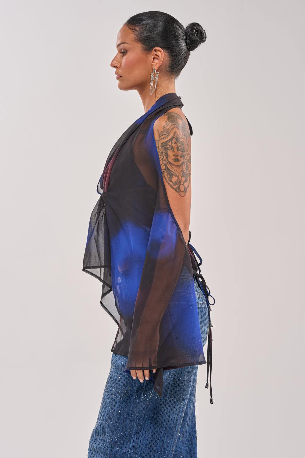 A woman with dark hair in a bun wears the FLY AWAY TIE FRONT BLOUSE in black and blue marbled chiffon, styled as a halter top with a denim skirt and hoop earrings, standing in profile to show her tattooed arm. 3