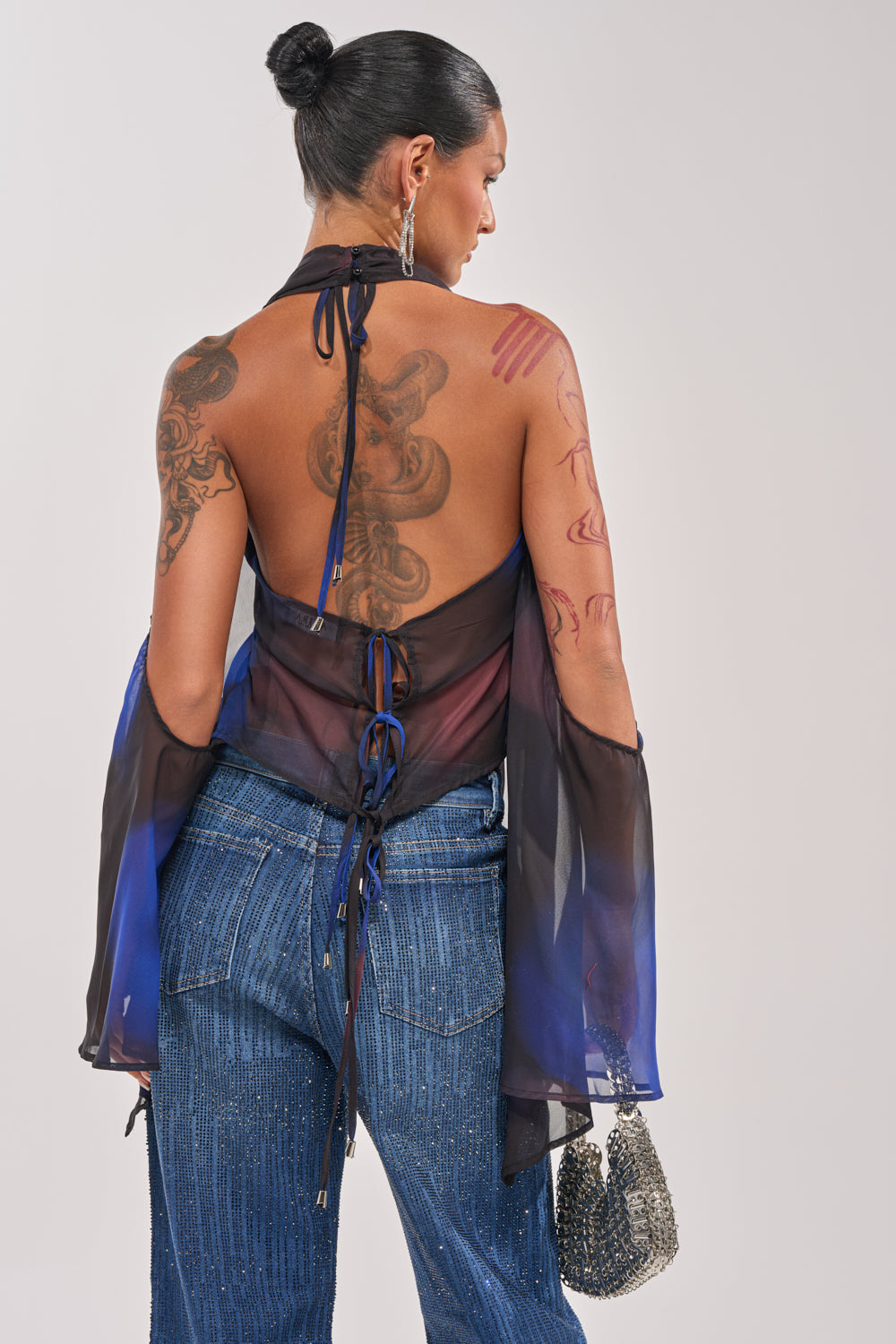 A tattooed woman with dark hair in a bun wears the FLY AWAY TIE FRONT BLOUSE—blue marbled chiffon with loose sleeves and open back—styled with sparkly blue jeans and a metallic handbag, facing away from the camera for a chic vacation look. 4
