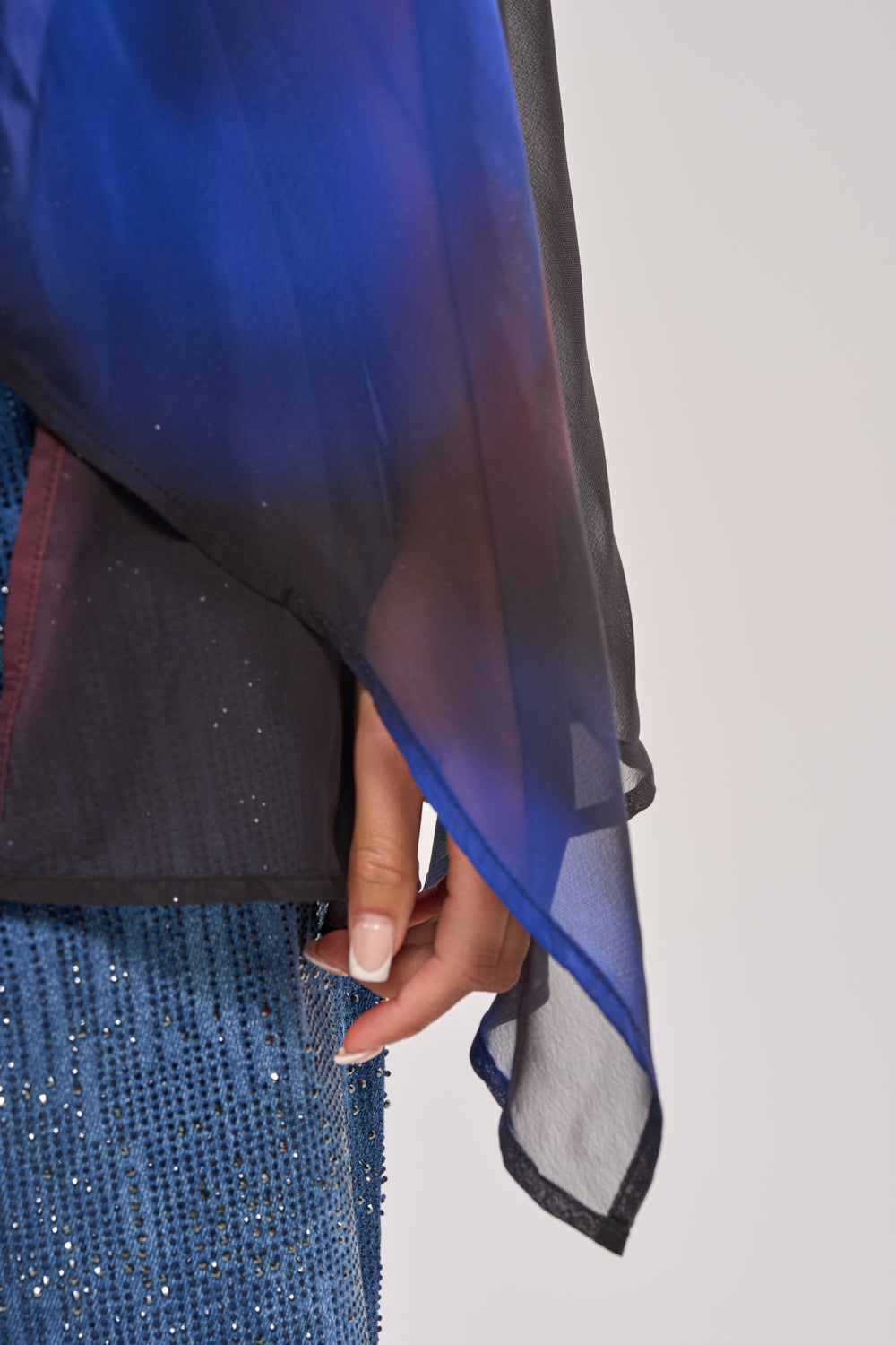 A close-up of a person's hand by their side, wearing the FLY AWAY TIE FRONT BLOUSE—a sheer blue marbled chiffon piece over a sparkling blue garment—standing out against a plain, light-colored background. 7