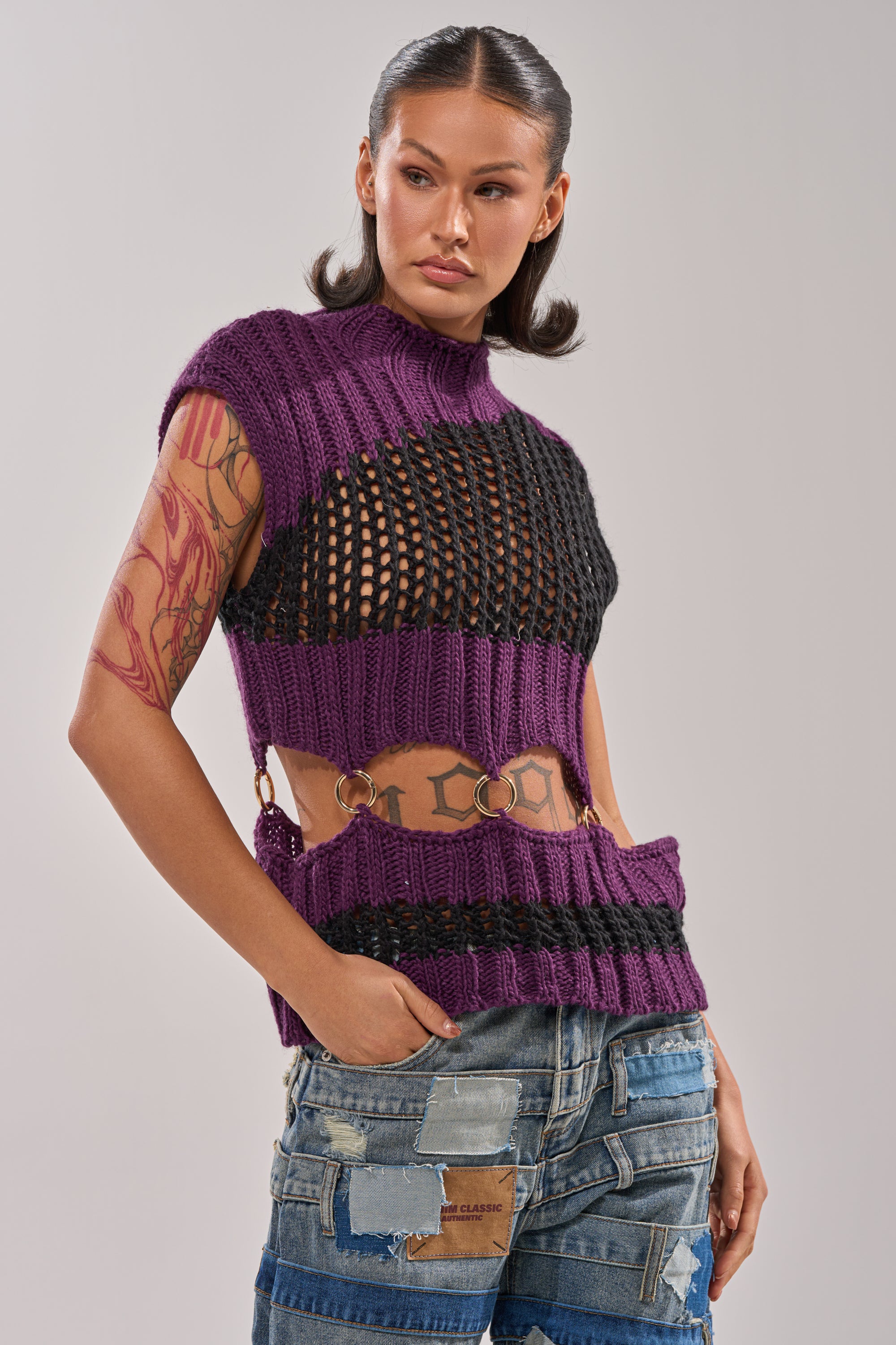 A woman with long dark hair and tattoos wears the LOUD AND WILD SWEATER in purple and black, featuring cut-outs and metal rings, paired with patchwork denim jeans. She stands with one hand in her pocket, looking to the side. 0