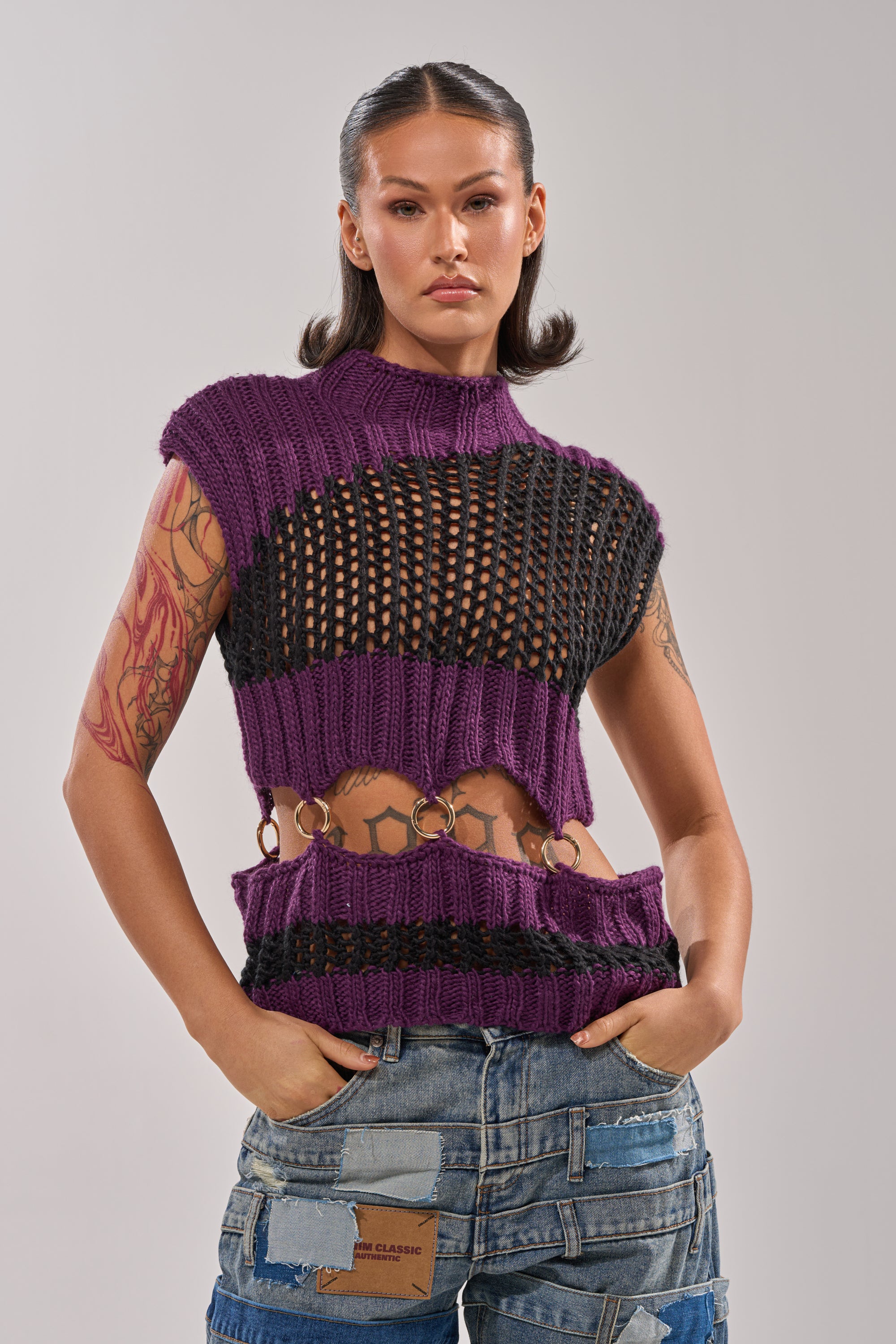 A woman with tattoos models the LOUD AND WILD SWEATER, a purple and black cut-out knit with metal rings, paired with patchwork denim jeans—an edgy outfit ideal for festivals. 1