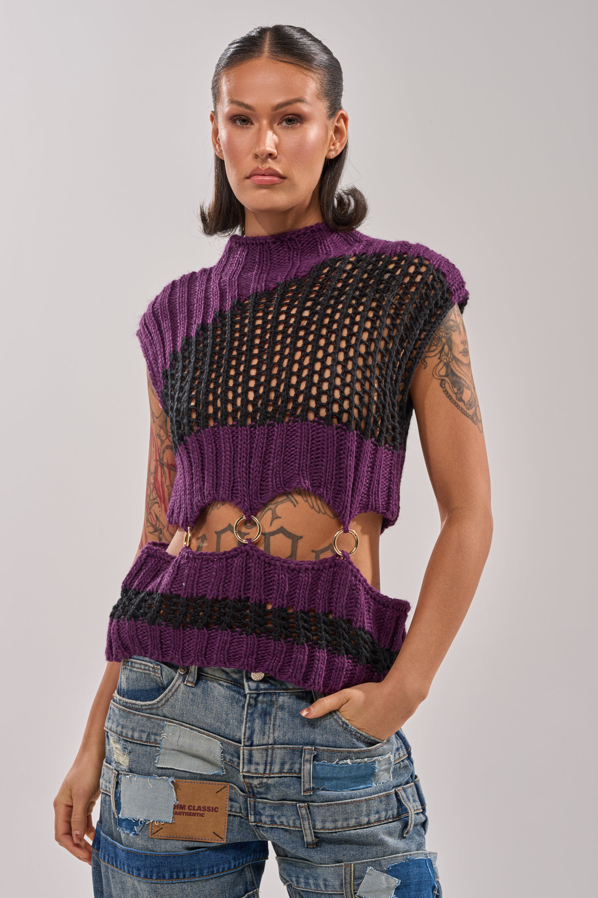 A woman stands against a plain background wearing the LOUD AND WILD SWEATER, a purple and black cut-out knit with gold hardware, paired with blue patchwork jeans and one hand in her pocket. 2
