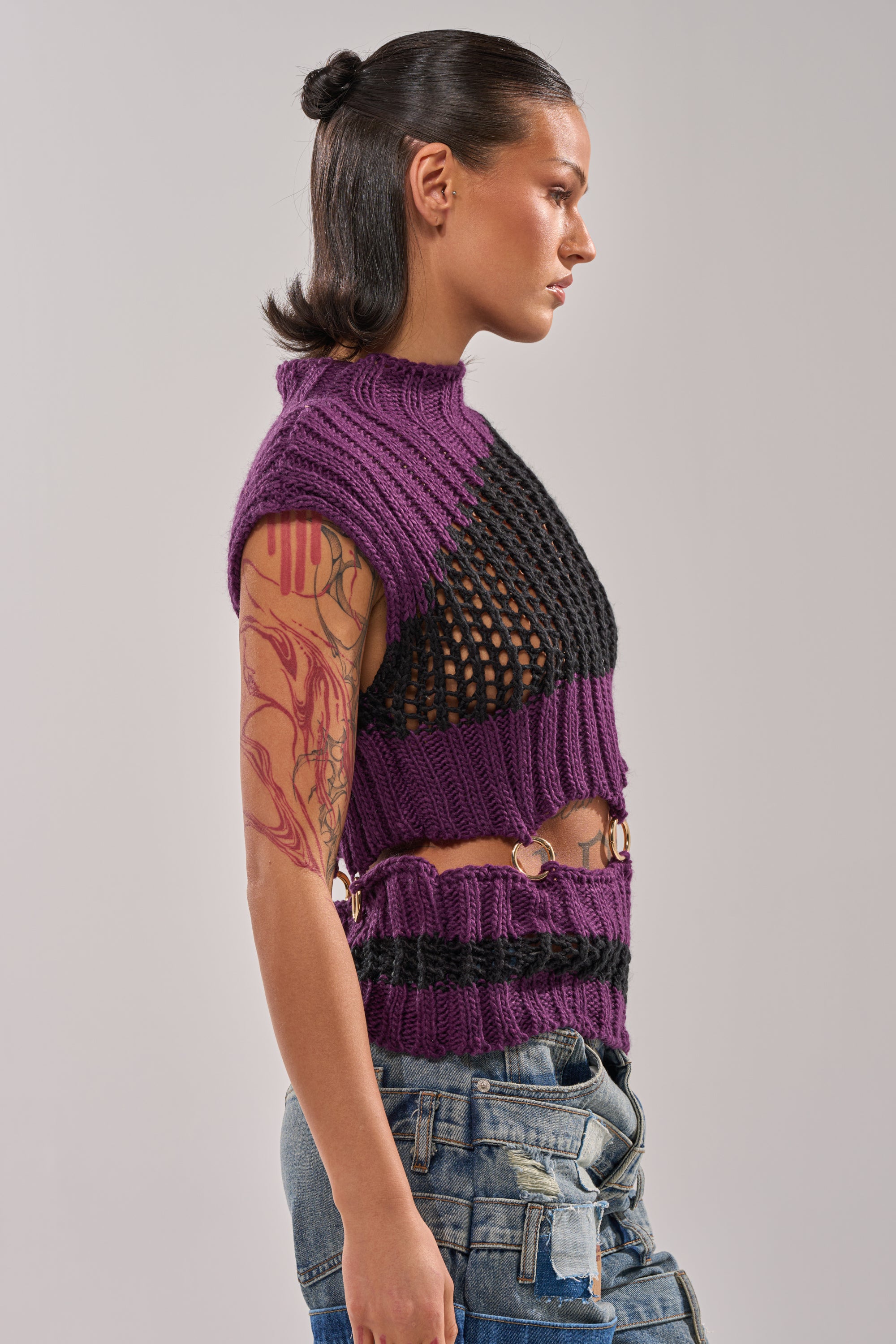 A person with dark hair styled back and a colorful tattoo sleeve on their left arm stands in profile against a plain background, wearing the LOUD AND WILD SWEATER—a purple and black cut-out knit with gold hardware—paired with distressed jeans. 3