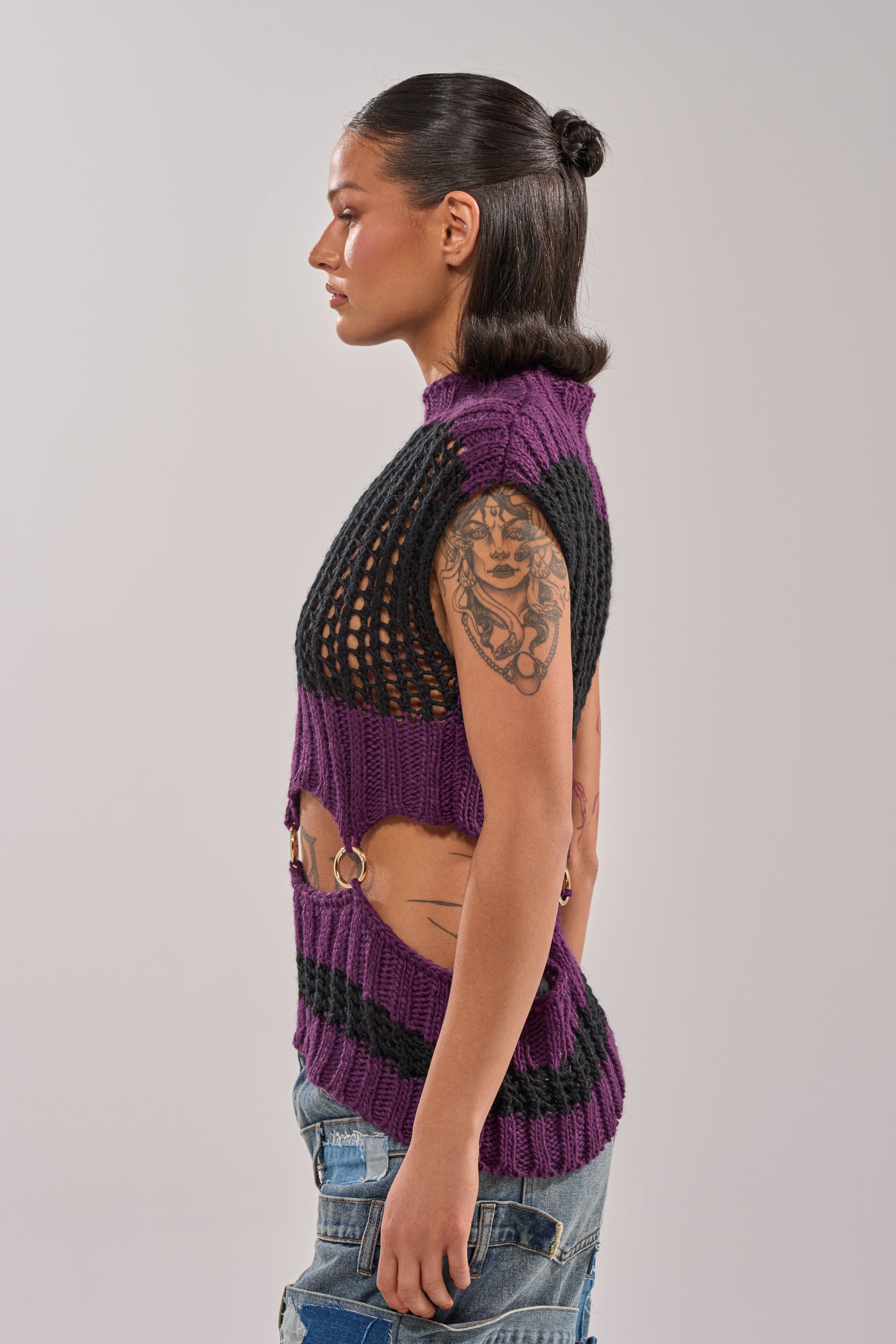 A person with dark hair in a bun stands in profile, wearing the LOUD AND WILD SWEATER—a sleeveless, purple and black striped knit with side cutouts and gold hardware—paired with distressed blue jeans. Their upper arm features a large tattoo. 4