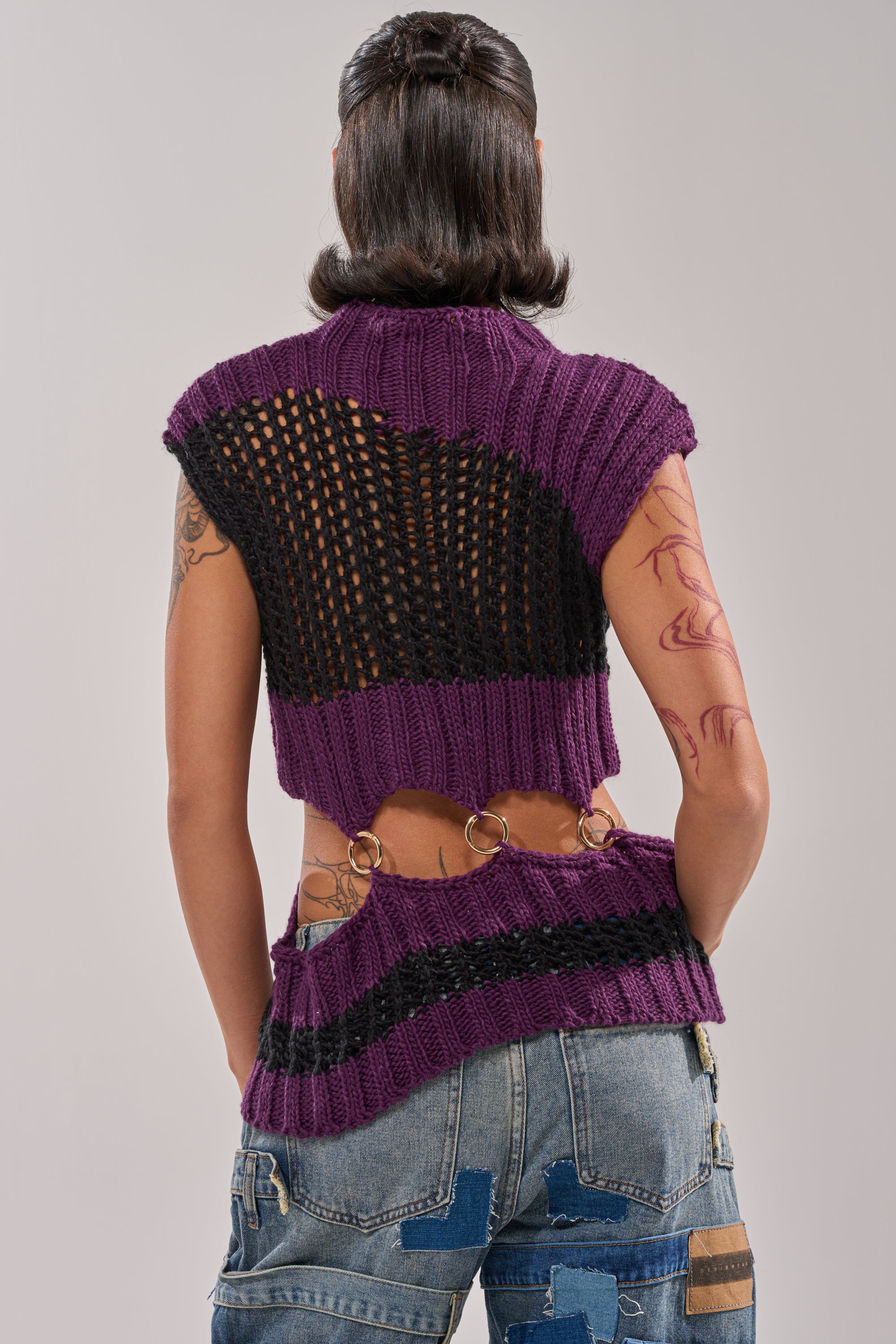 A person stands facing away in streetwear: the LOUD AND WILD SWEATER, a cut-out knit with purple and black details plus metal rings. Tattoos cover their arms, and they finish the look with patchwork blue jeans. 5