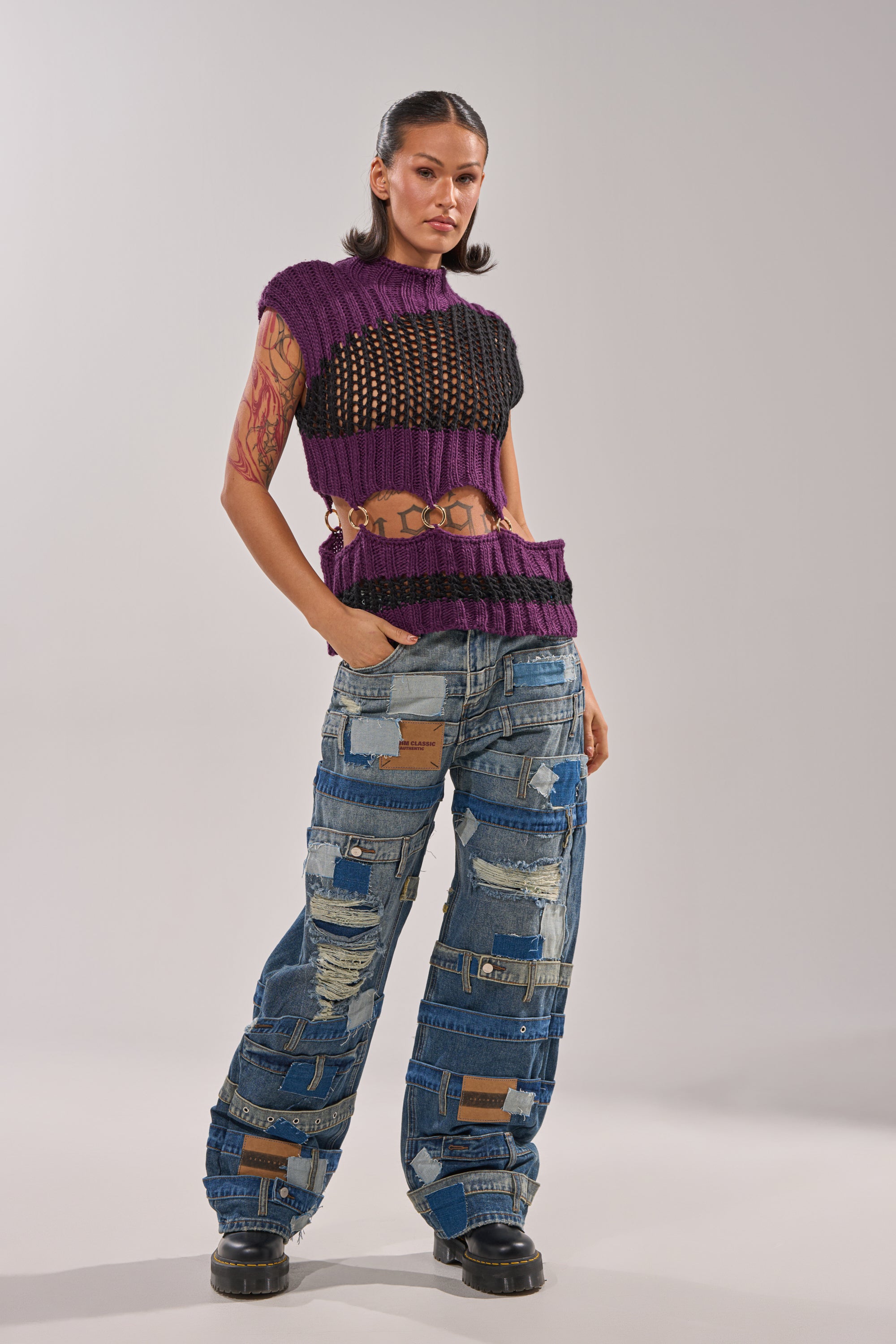A woman exudes confidence in the LOUD AND WILD SWEATER, a sleeveless purple and black knit crop top, paired with wide-leg patchwork jeans. She sports dark slicked-back hair, visible tattoos, and black boots for a bold streetwear look. 6