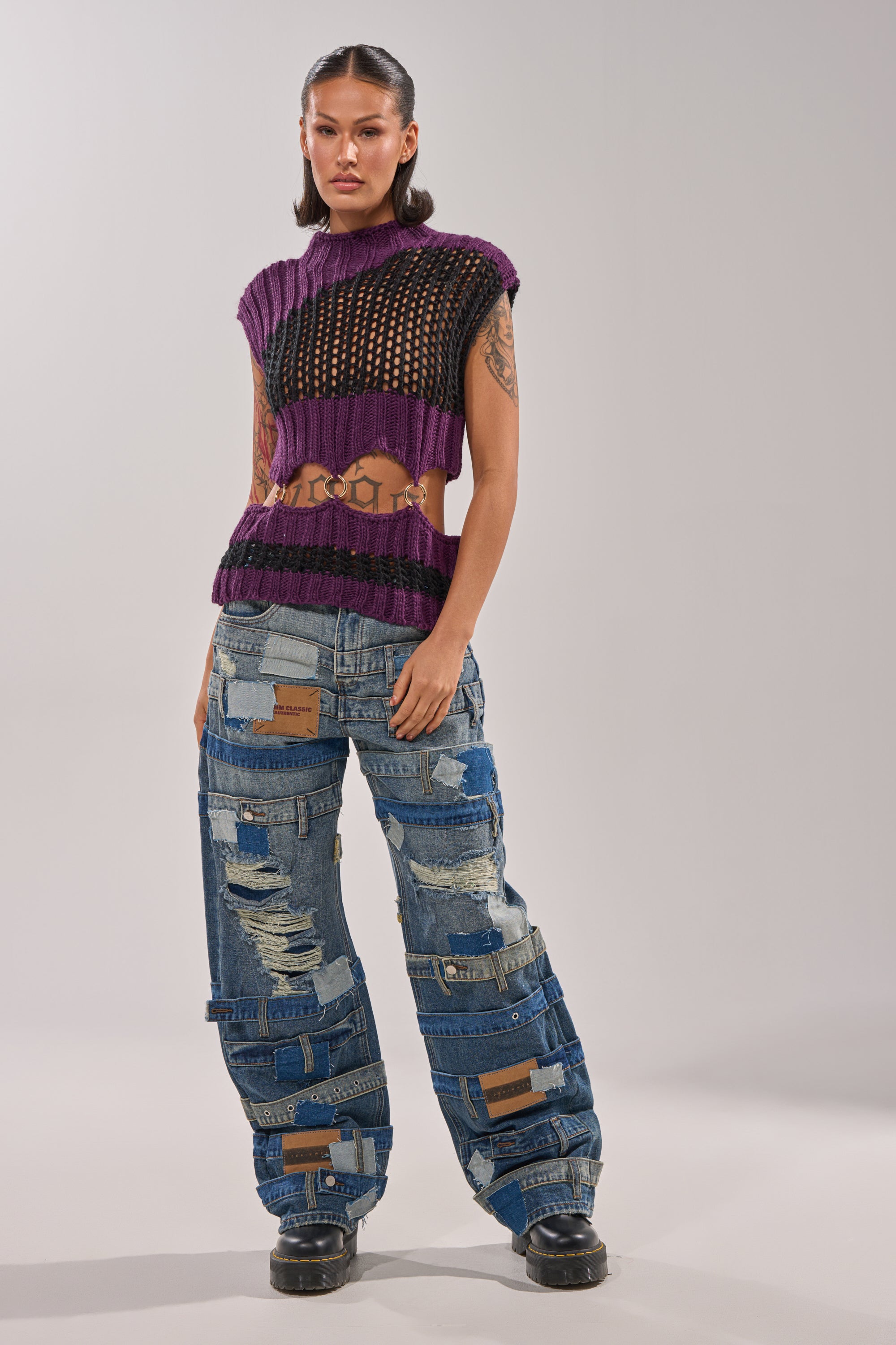A woman stands confidently in the LOUD AND WILD SWEATER—a purple cut-out knit—paired with high-waisted patchwork jeans, black platform shoes, and visible tattoos, channeling bold streetwear style against a plain background. 7