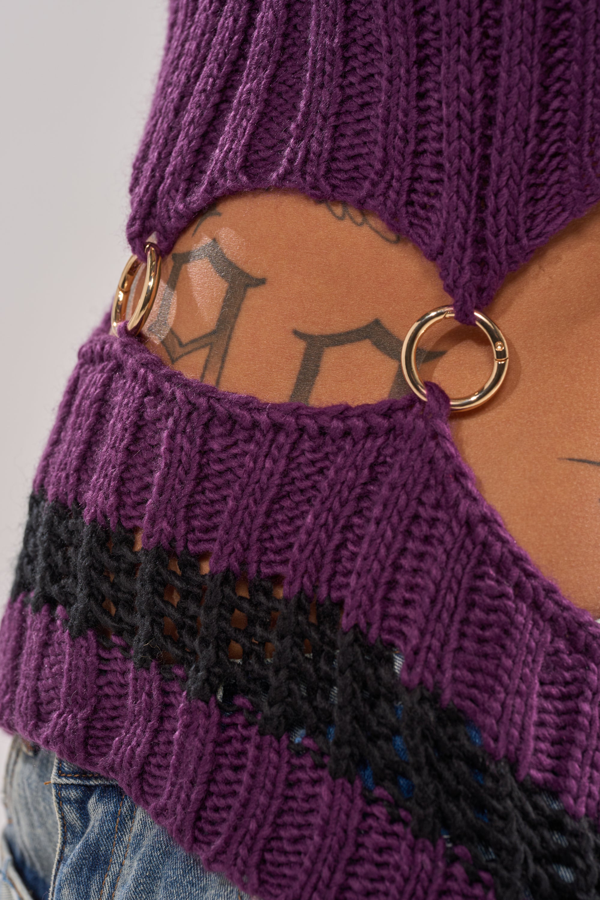 Close-up of a person's lower back in the LOUD AND WILD SWEATER, a purple cut-out knit with gold rings linking the fabric. Black tattoos show on their skin, paired with blue denim jeans for a streetwear vibe. 8