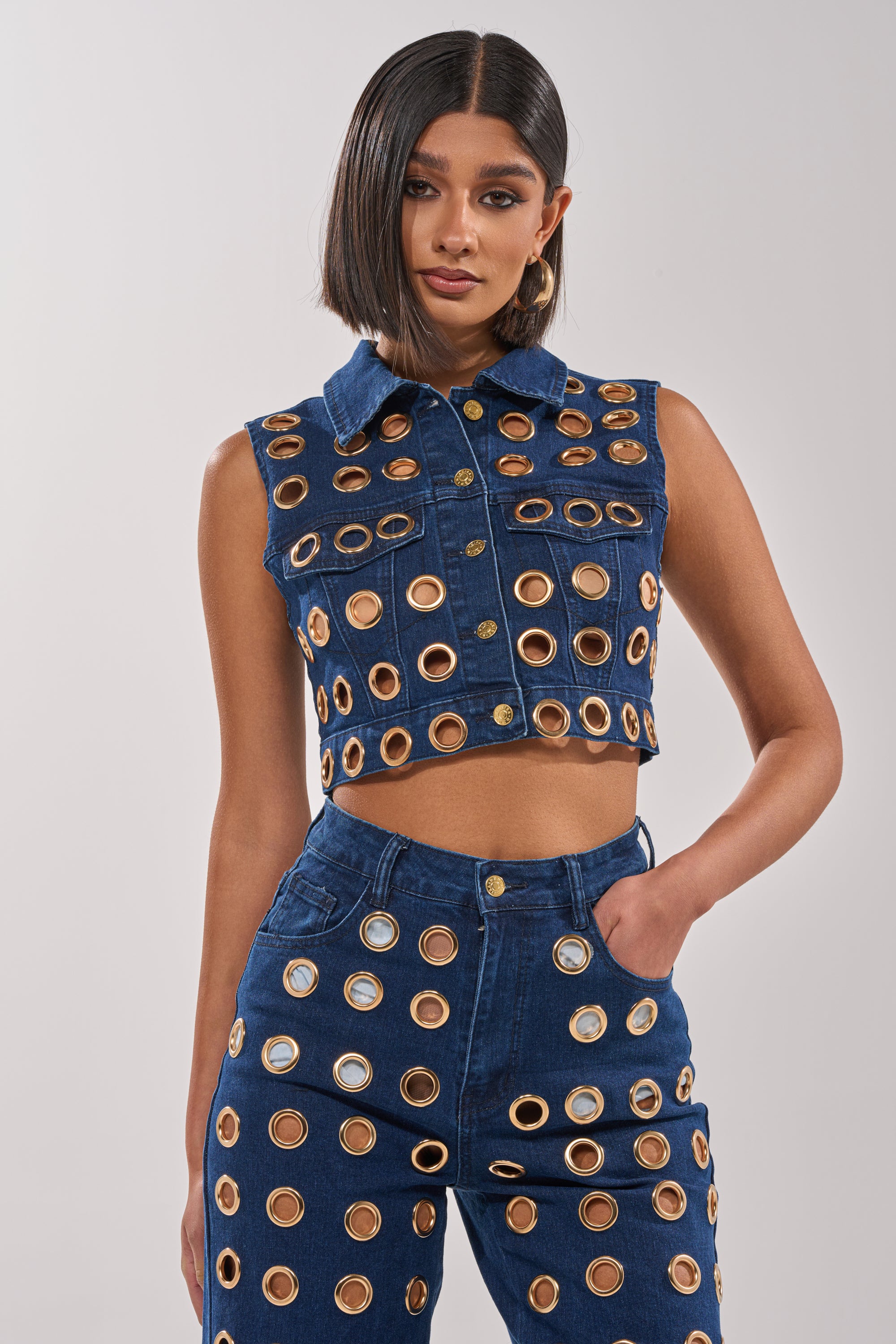 A woman models the SEE THROUGH ME BUTTON DOWN with high-waisted pants featuring metallic grommets, creating a bold streetwear look. Her straight, shoulder-length hair and relaxed pose complete this festival-ready style against a plain background.