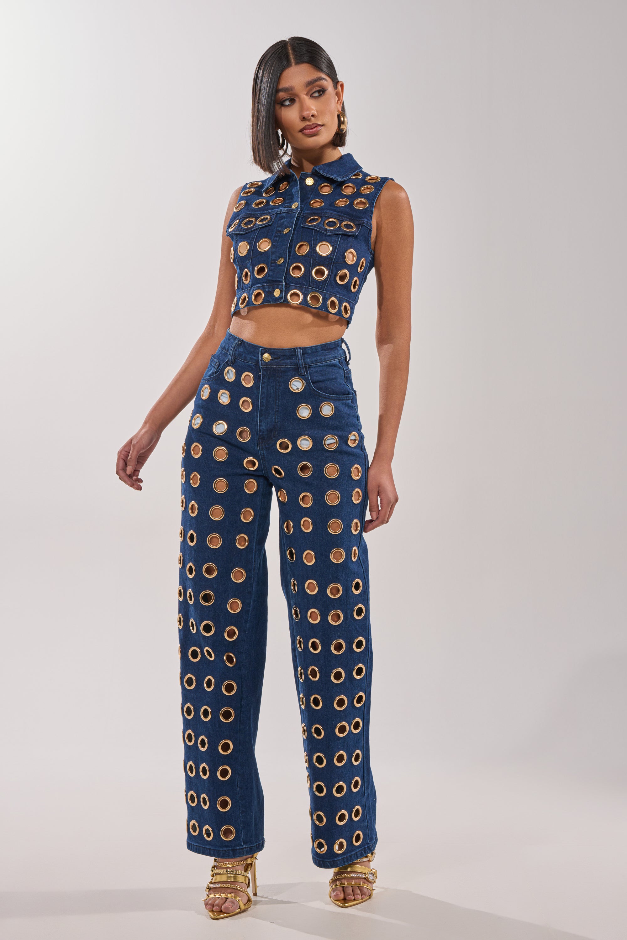 A woman stands confidently in gold heels against a light gray background, wearing the SEE THROUGH ME WIDE LEG DENIM with bold gold grommet accents and a sleeveless, cropped denim vest. 1