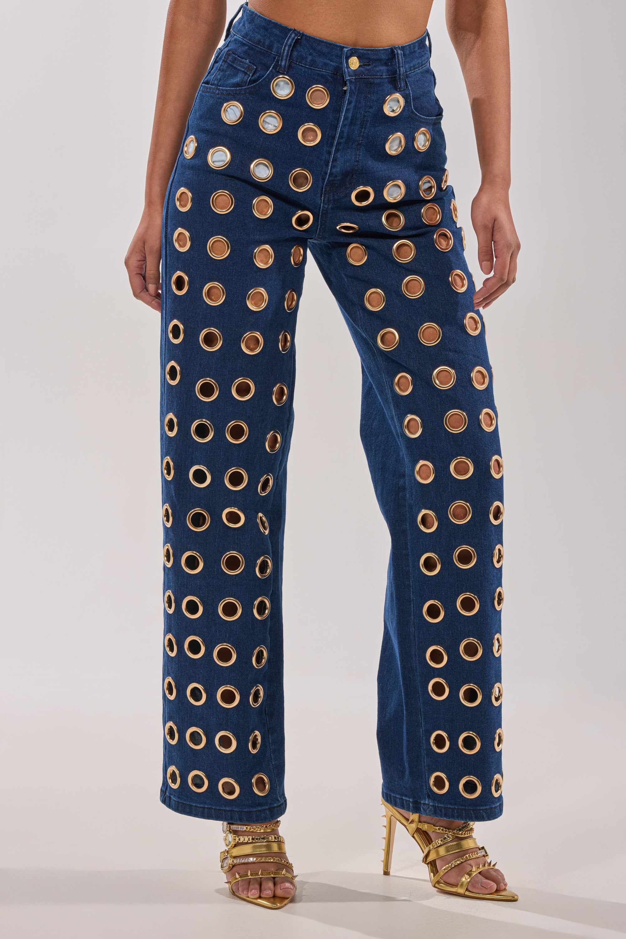 A person wears SEE THROUGH ME WIDE LEG DENIM, high-waisted jeans adorned with gold metal grommets and featuring a wide-leg fit, paired with strappy gold high-heeled sandals for a striking, statement look. 0