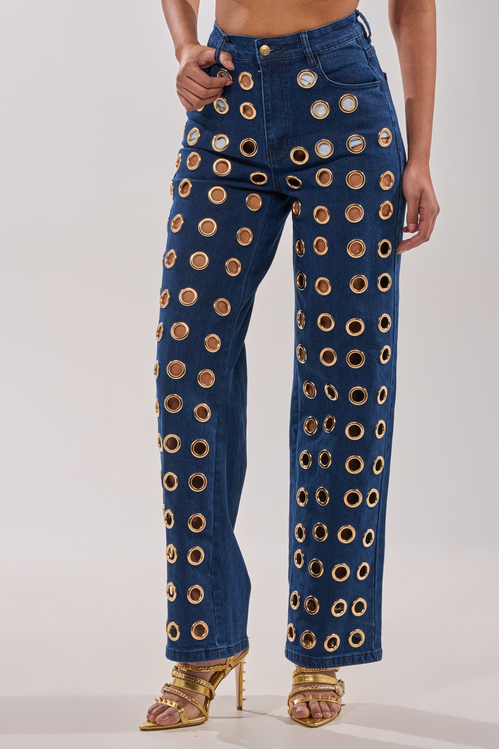 A person wears SEE THROUGH ME WIDE LEG DENIM with large metallic grommets, styled with gold strappy high-heeled sandals against a plain, light-colored background. 2
