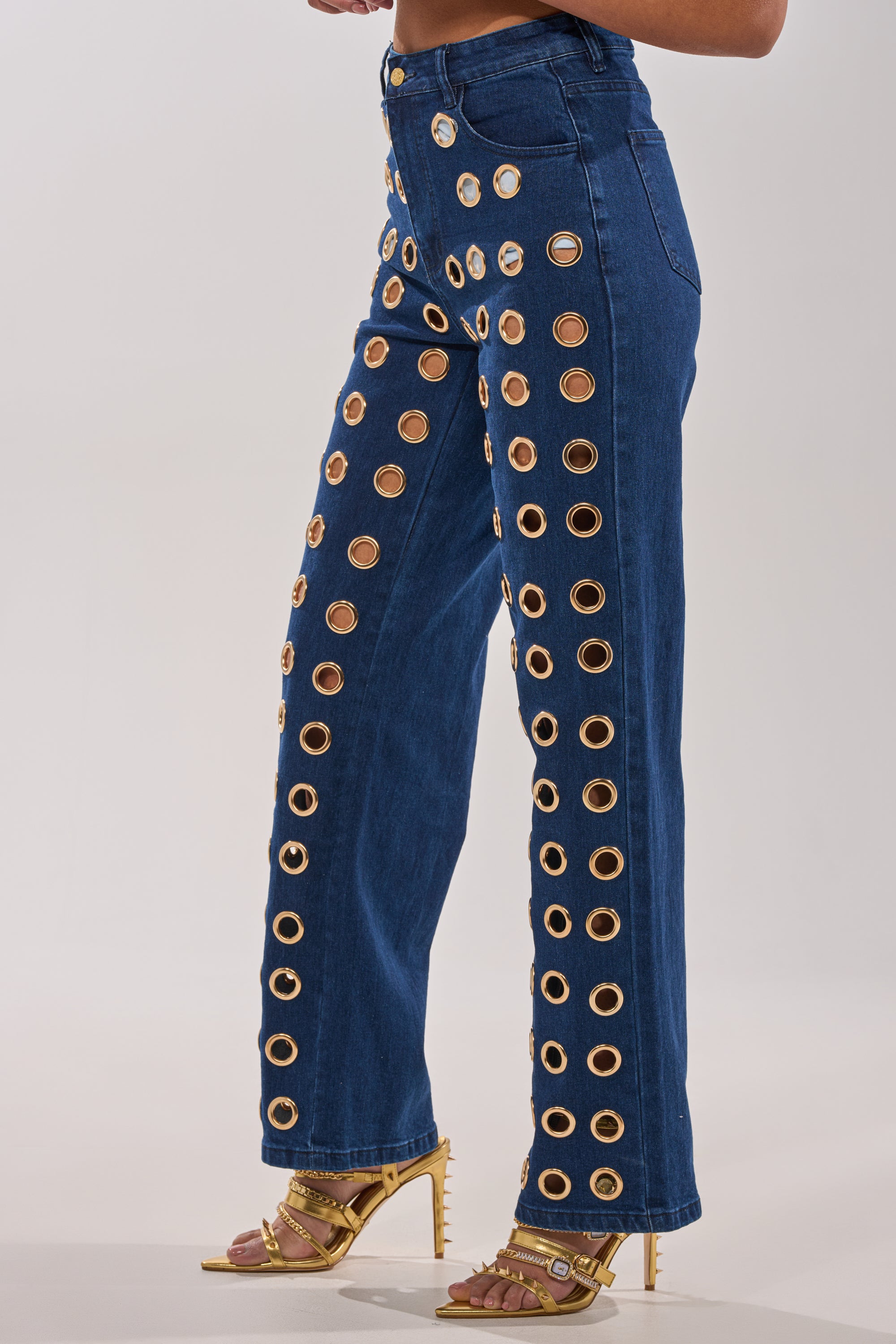 Wearing SEE THROUGH ME WIDE LEG DENIM, featuring high waist and bold gold grommets in vertical rows, paired with strappy gold heels. Only the torso and legs are visible against a plain background. 3