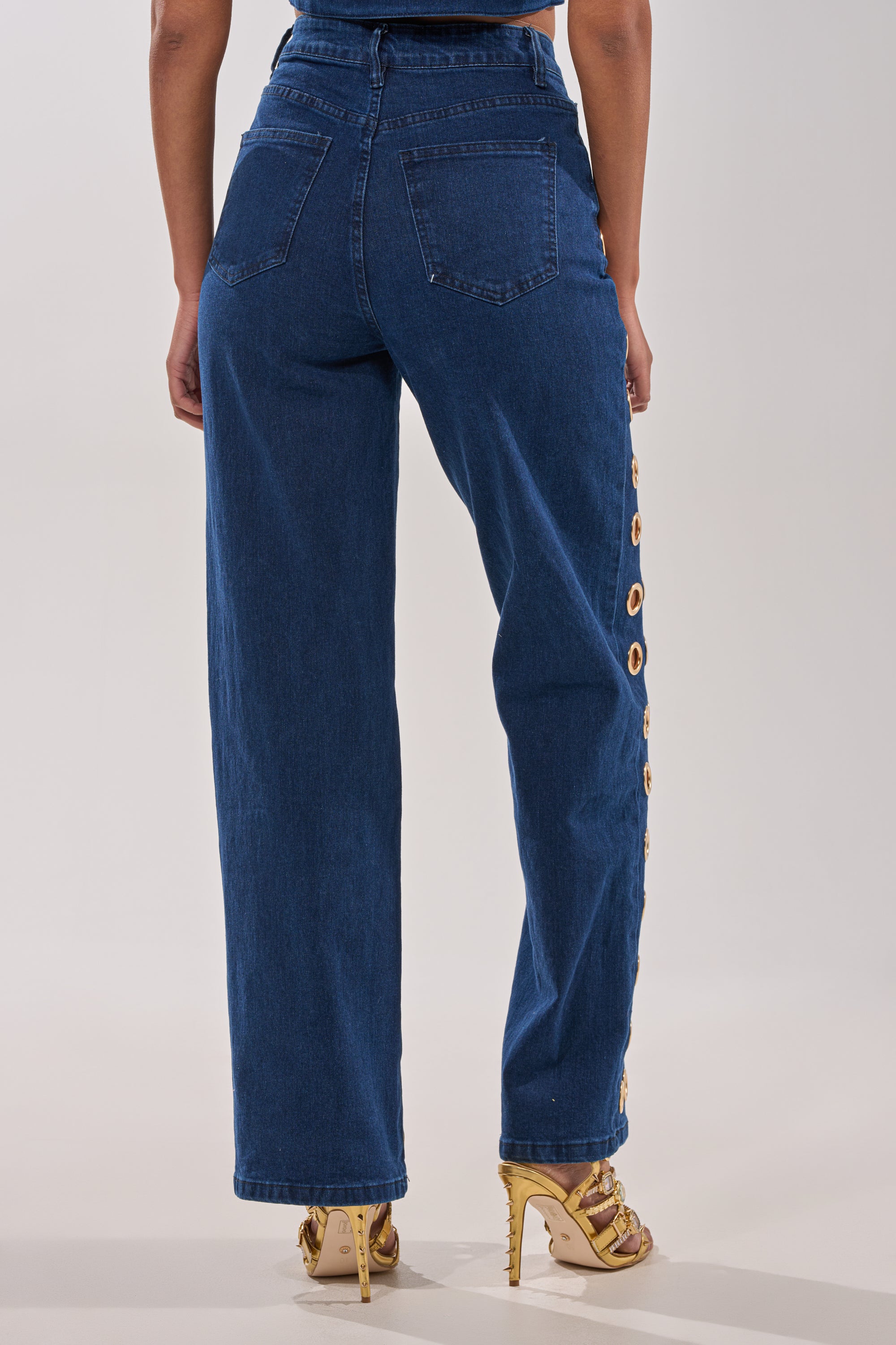 A person is seen from the back in SEE THROUGH ME WIDE LEG DENIM with side button details and gold high-heeled sandals against a plain light background. 4