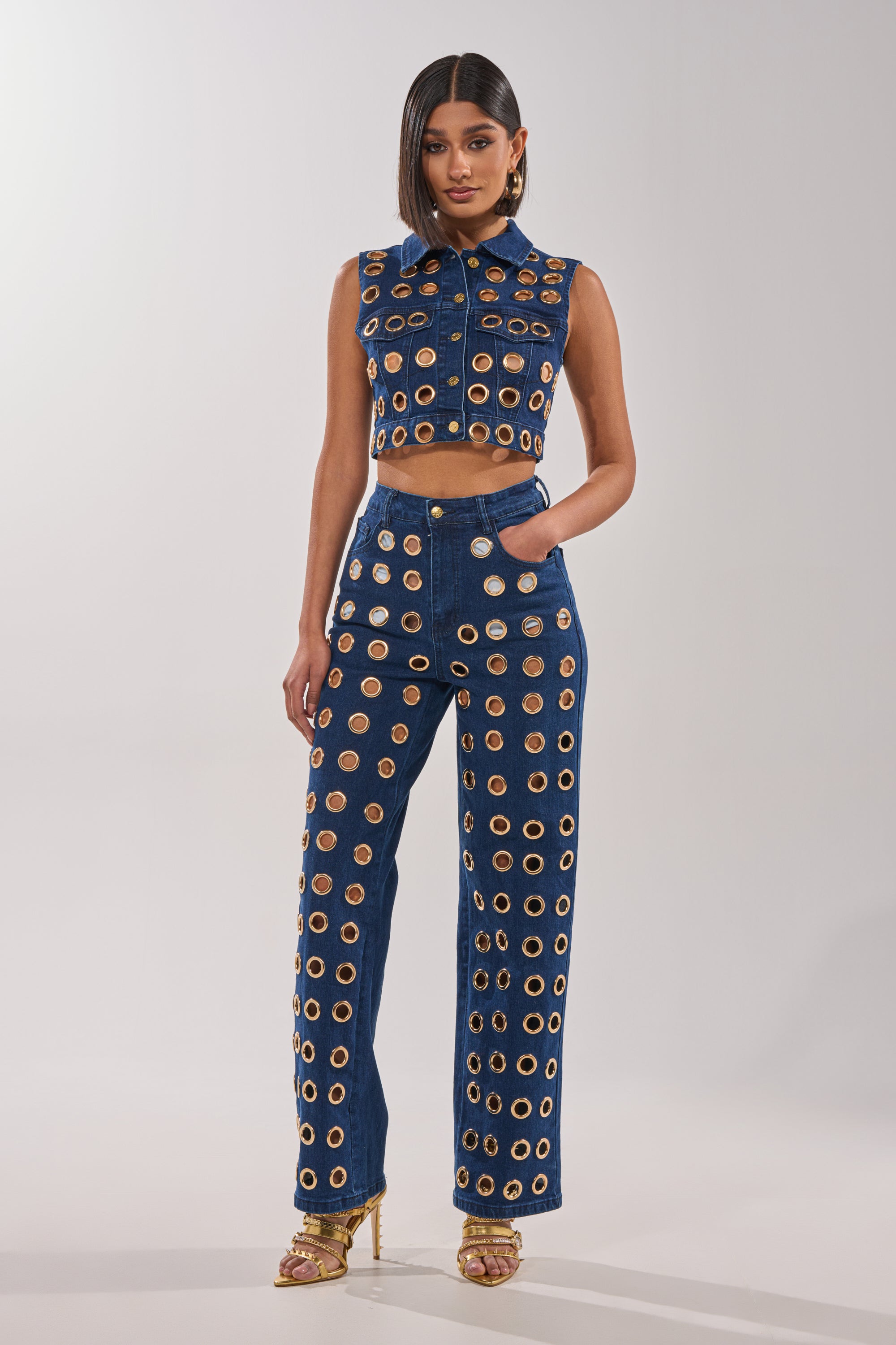 A woman poses confidently in SEE THROUGH ME WIDE LEG DENIM—a sleeveless, cropped denim top and high-waisted jeans with evenly spaced circular cutouts—styled with gold heels and statement earrings against a simple, light background. 5